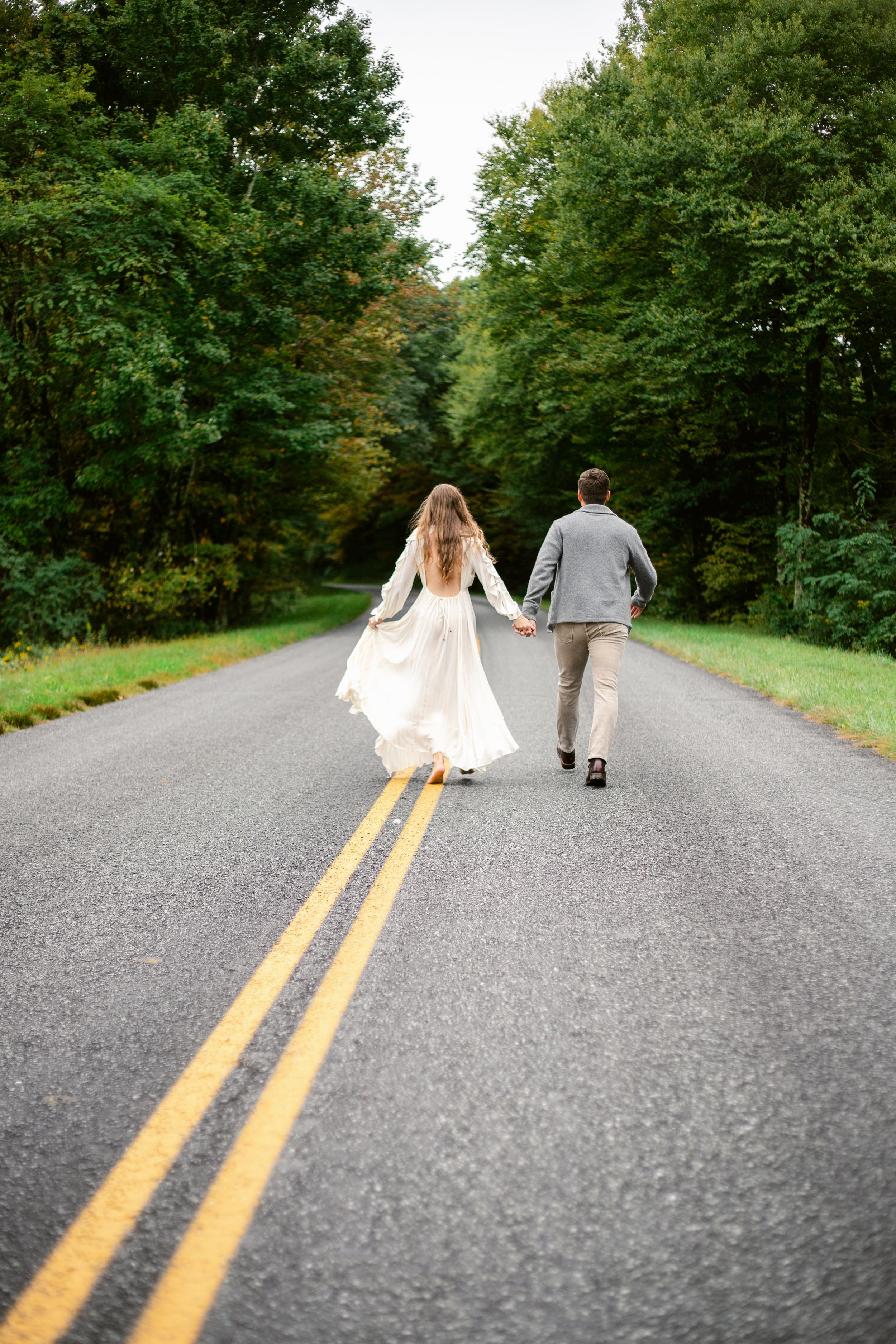 Couple walking down a tree-lined country road holding hands with the bride wearing a flowing white dress.