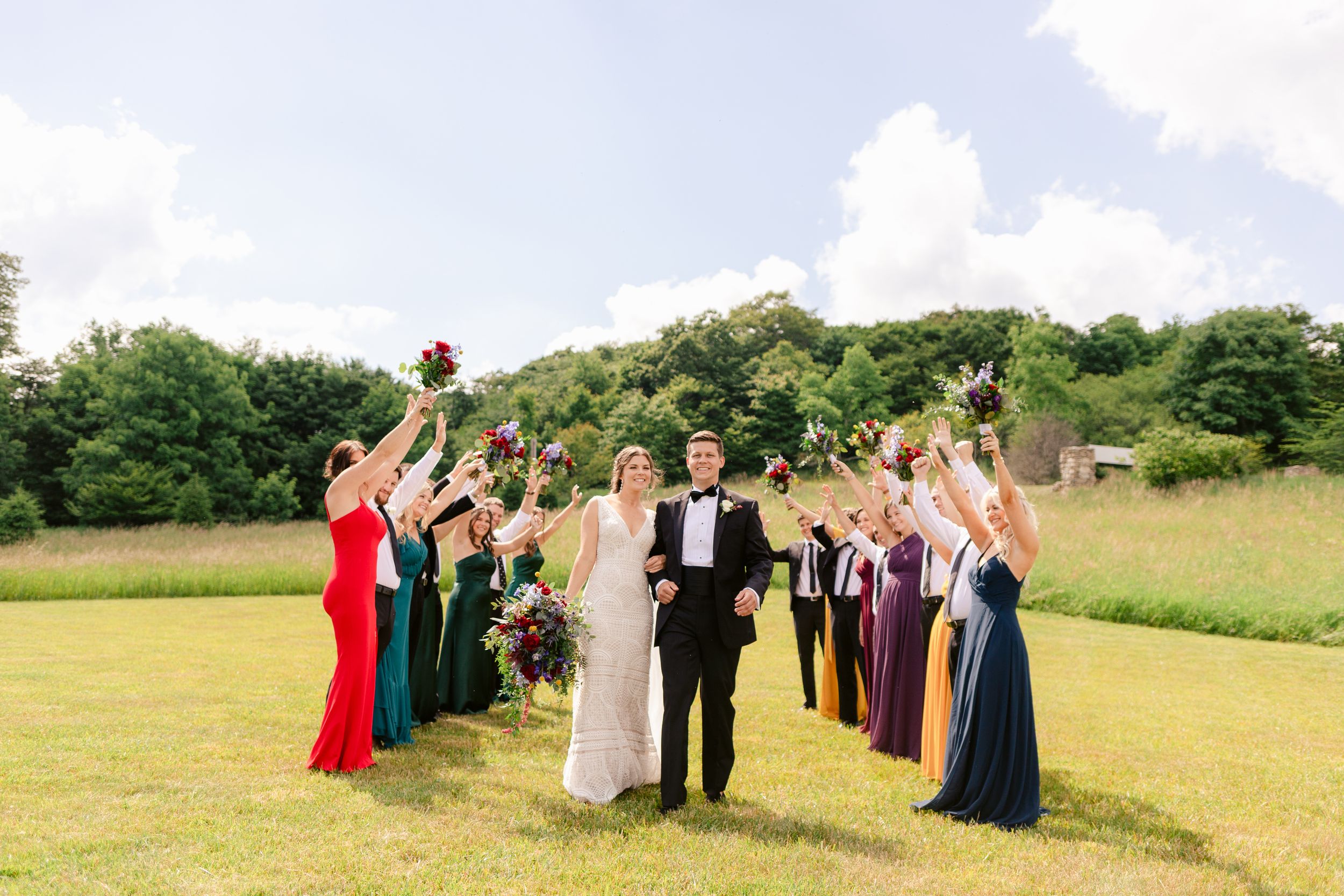 Joyful wedding party celebrating in sunny field with arms raised.