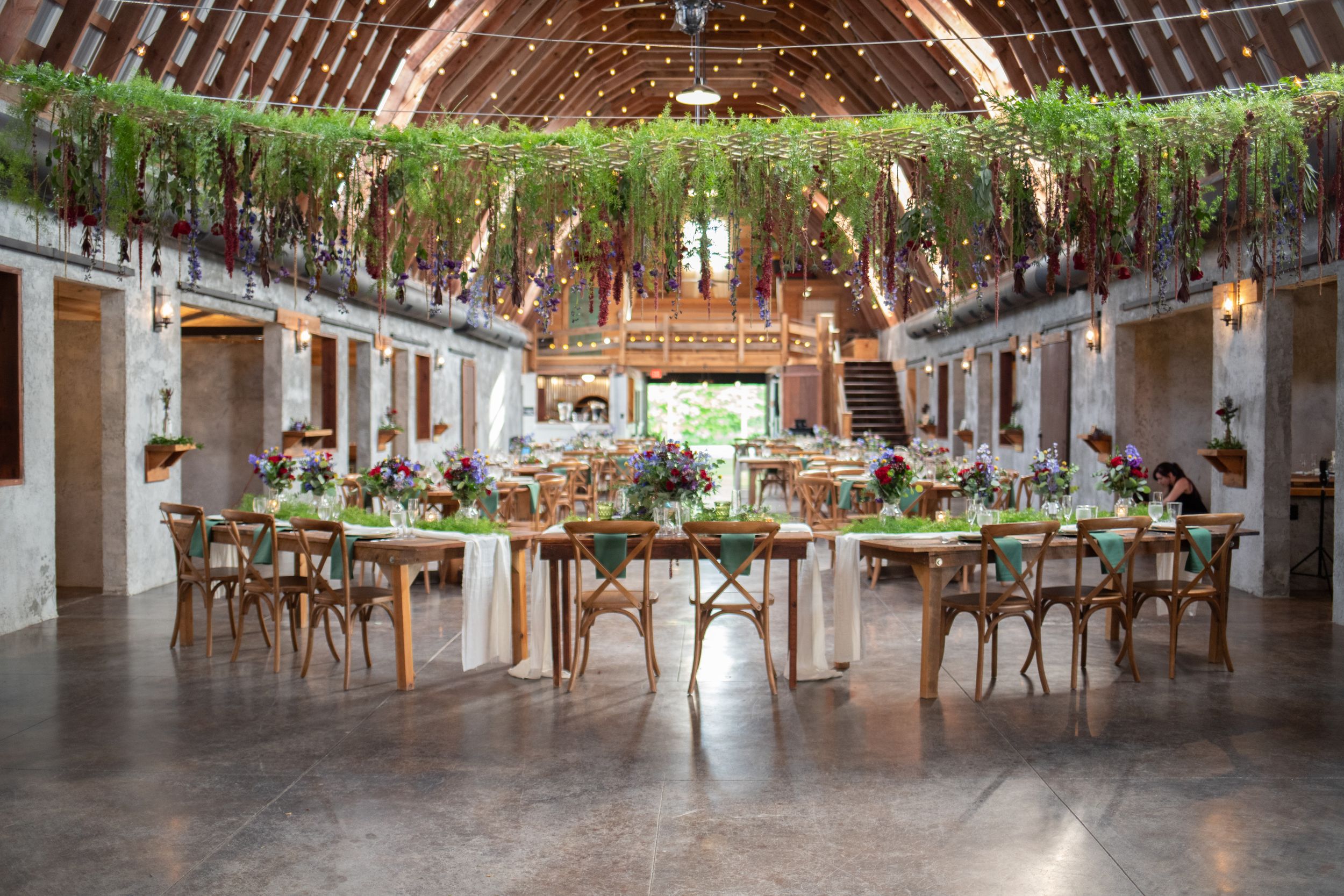 Rustic barn reception venue with vaulted ceiling, hanging greenery, and cross-back chairs.