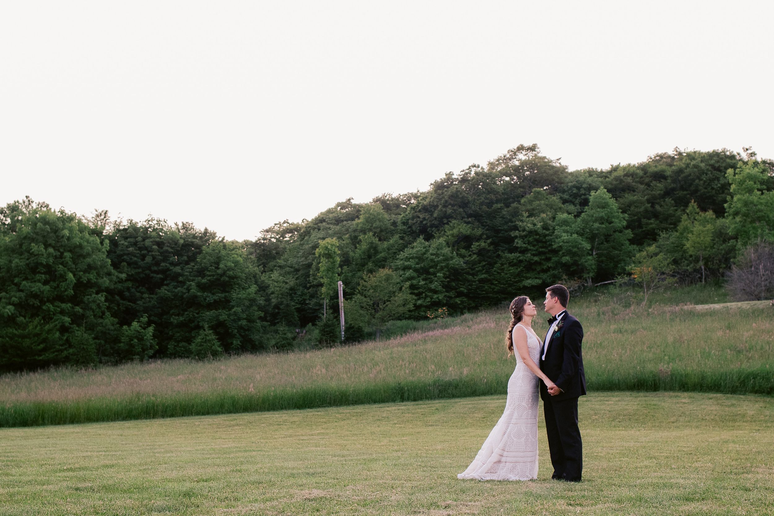Wedding couple stands in open field with tree line and sunset backdrop.