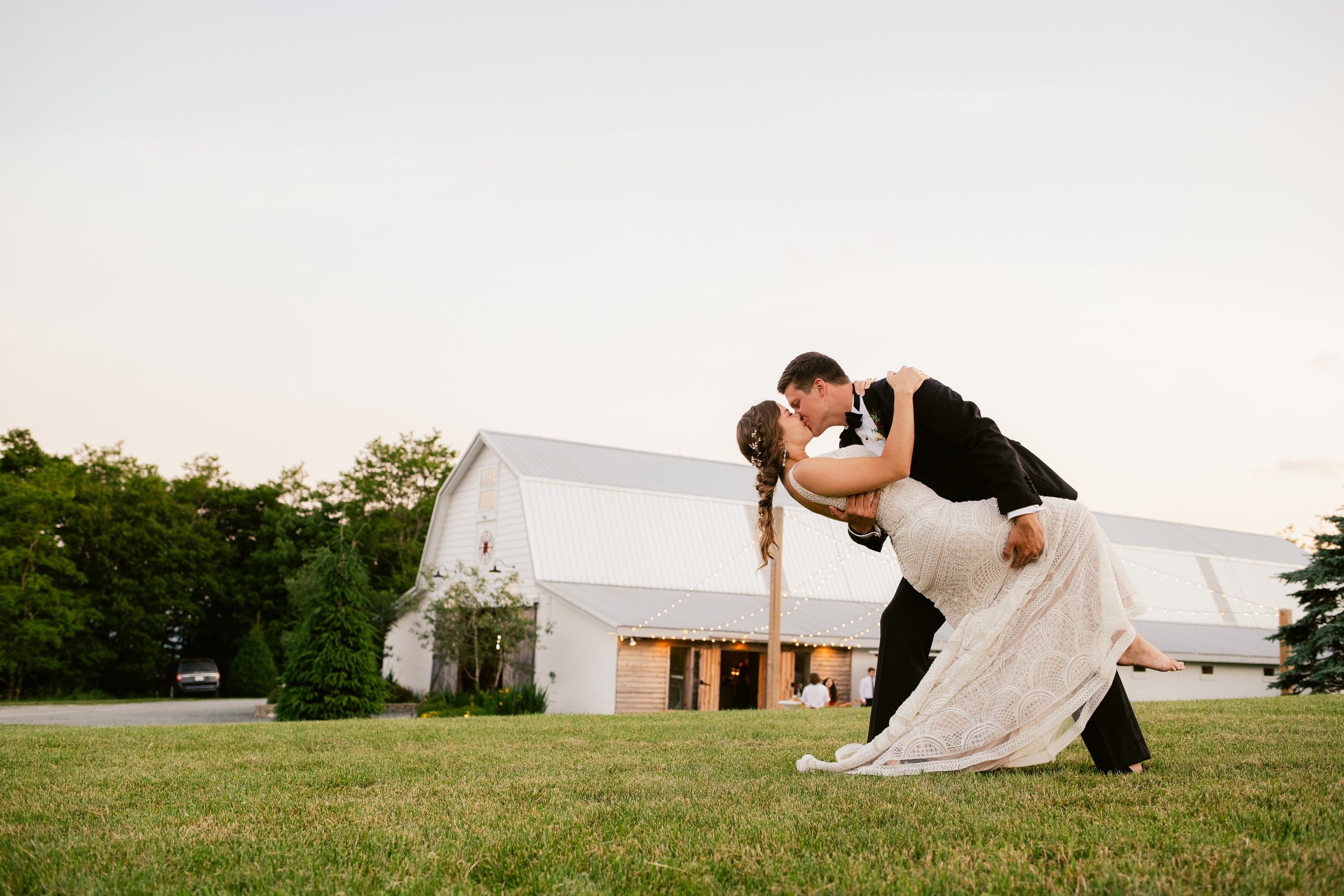 Romantic dip pose with white barn in background during sunset wedding photoshoot.