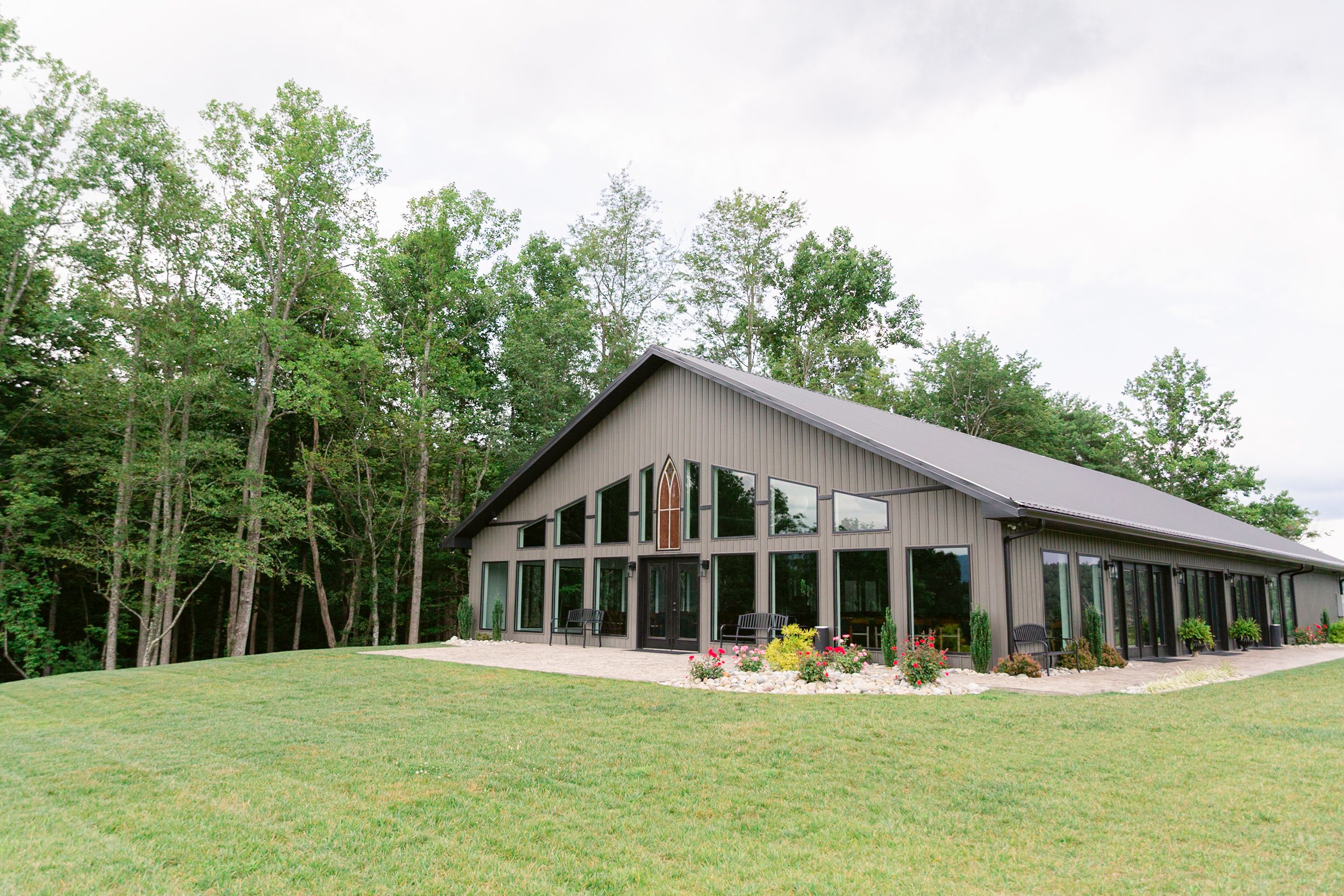 Modern barn-style venue with large windows and glass doors nestled among trees on a grassy lawn.