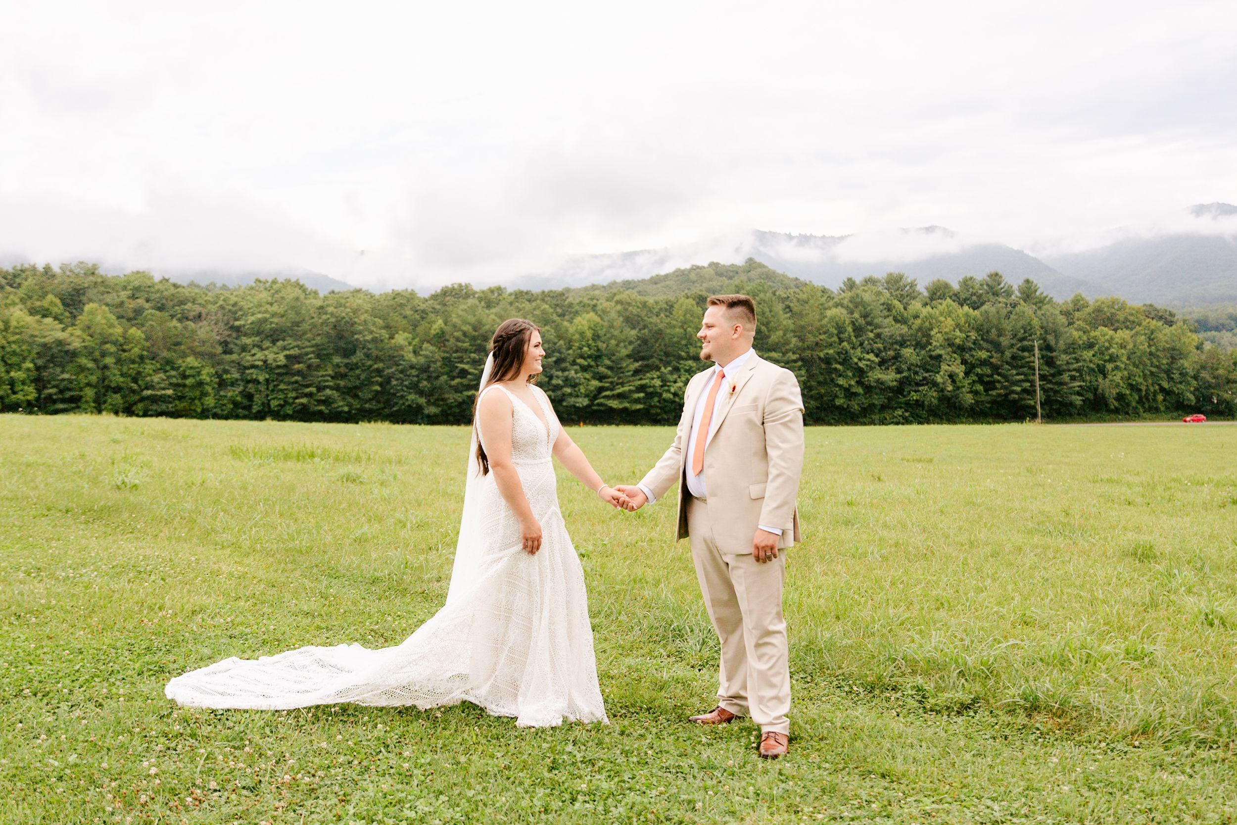A romantic couple walks together across a scenic hilltop meadow with mountain views in the background.