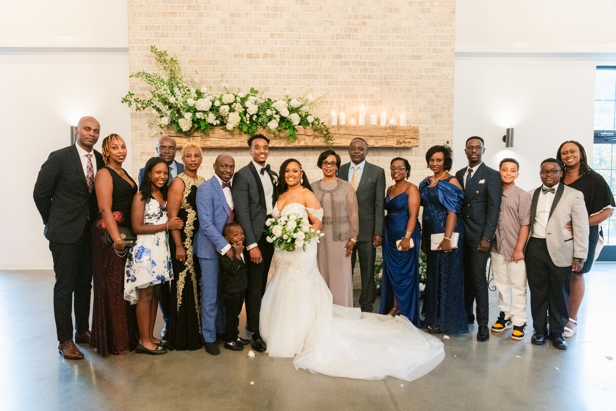 Wedding party group photo in front of white brick wall with floral arrangement at modern venue.