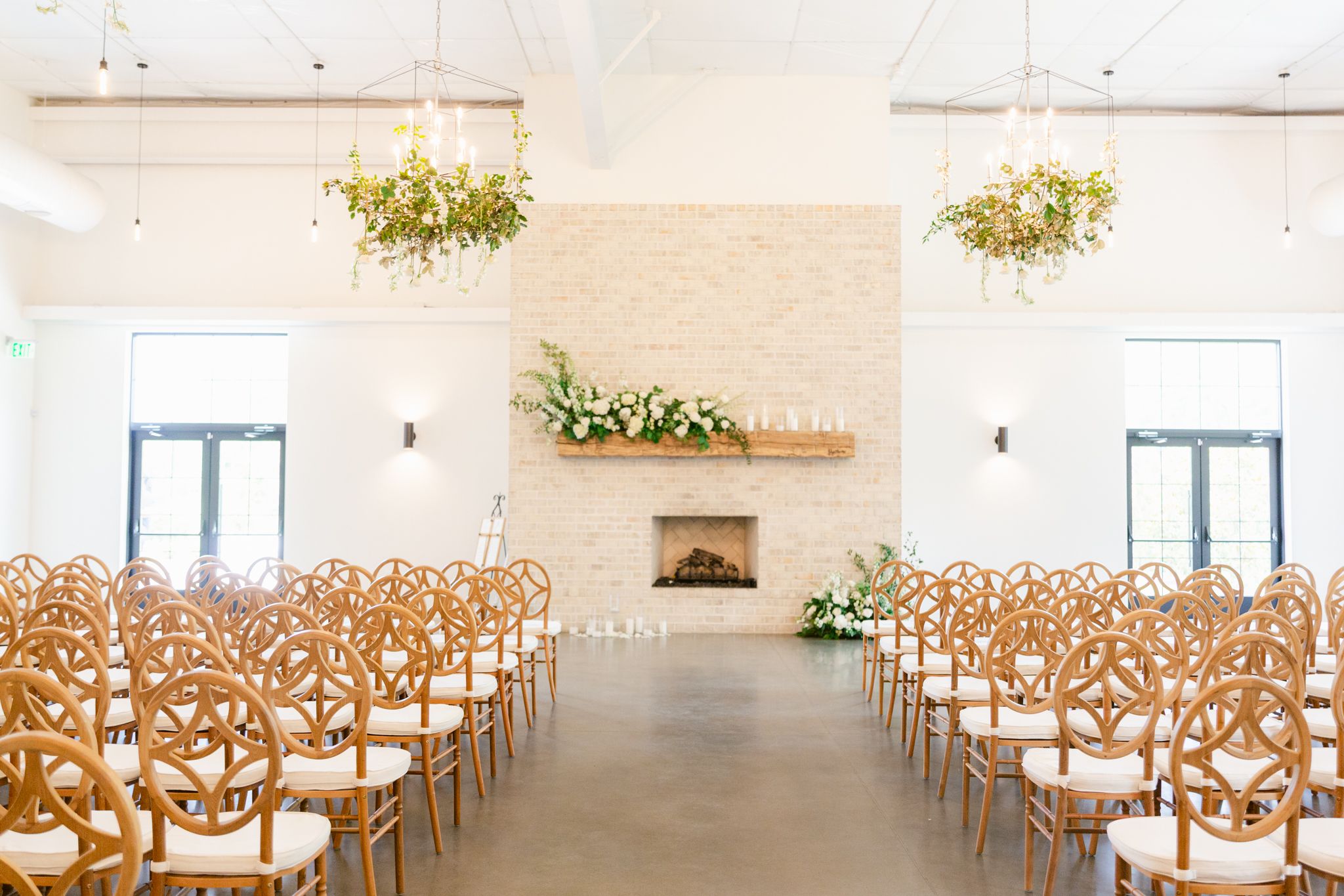Elegant white wedding ceremony venue with wooden chairs and hanging floral arrangements in a bright modern space.