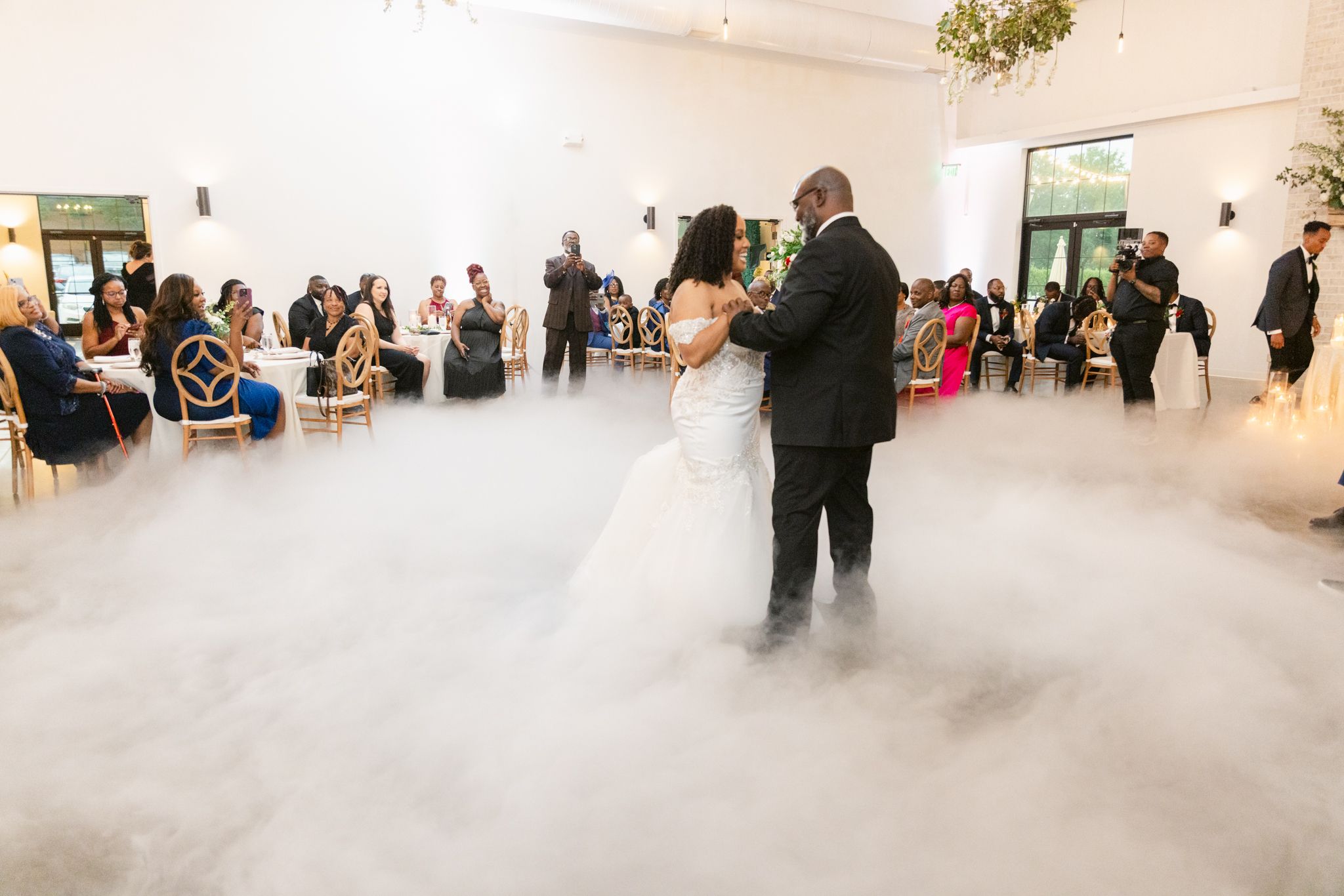 Wedding guests dancing and celebrating in an elegant white reception venue with high ceilings.