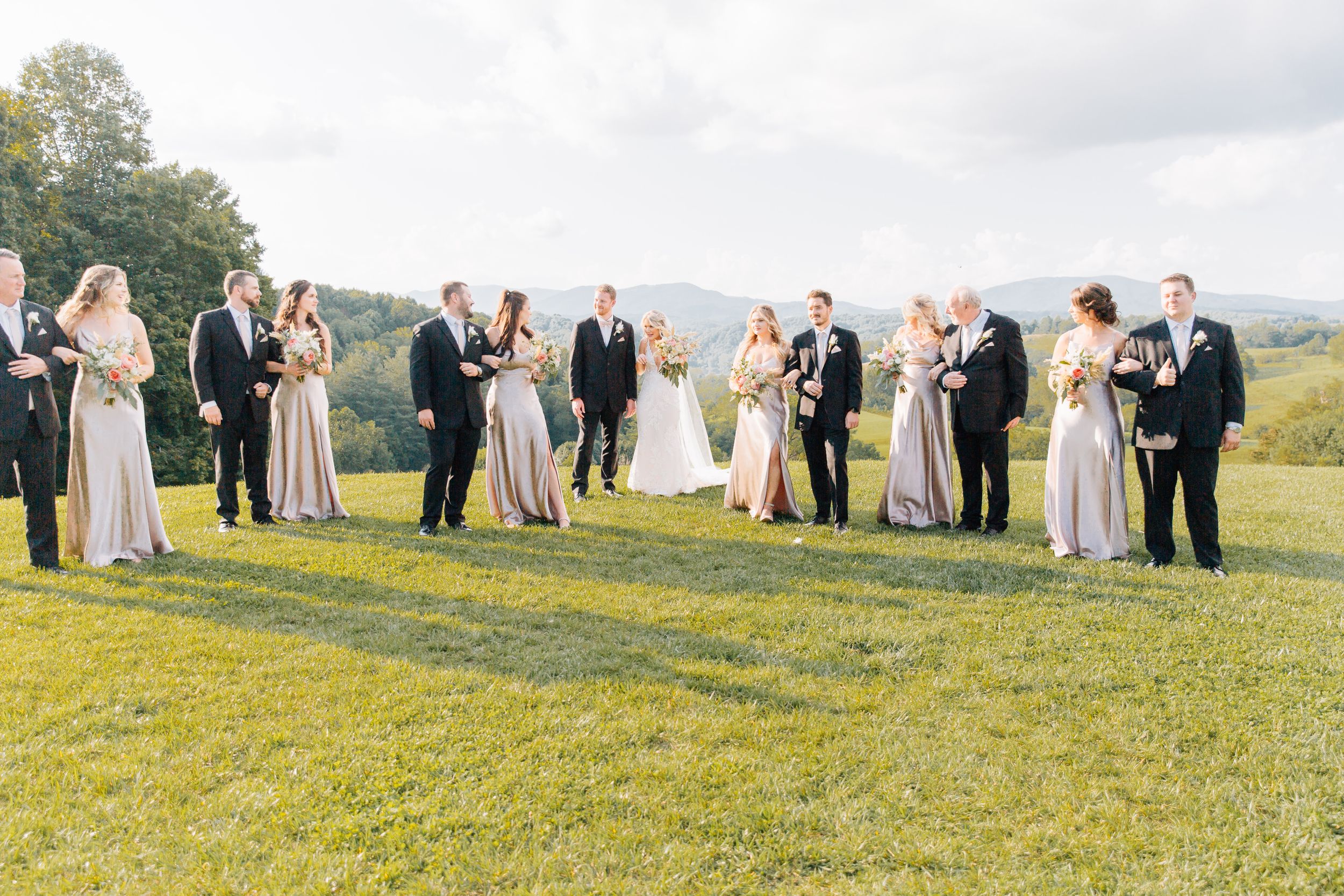 Wedding party poses on grassy hillside during golden hour with mountain views in background.