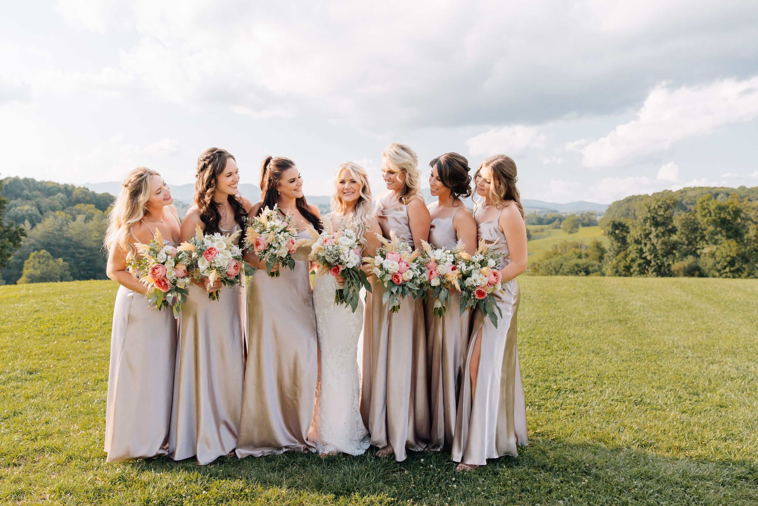 Bridesmaids in champagne dresses hold bouquets while standing in line on grassy hillside.