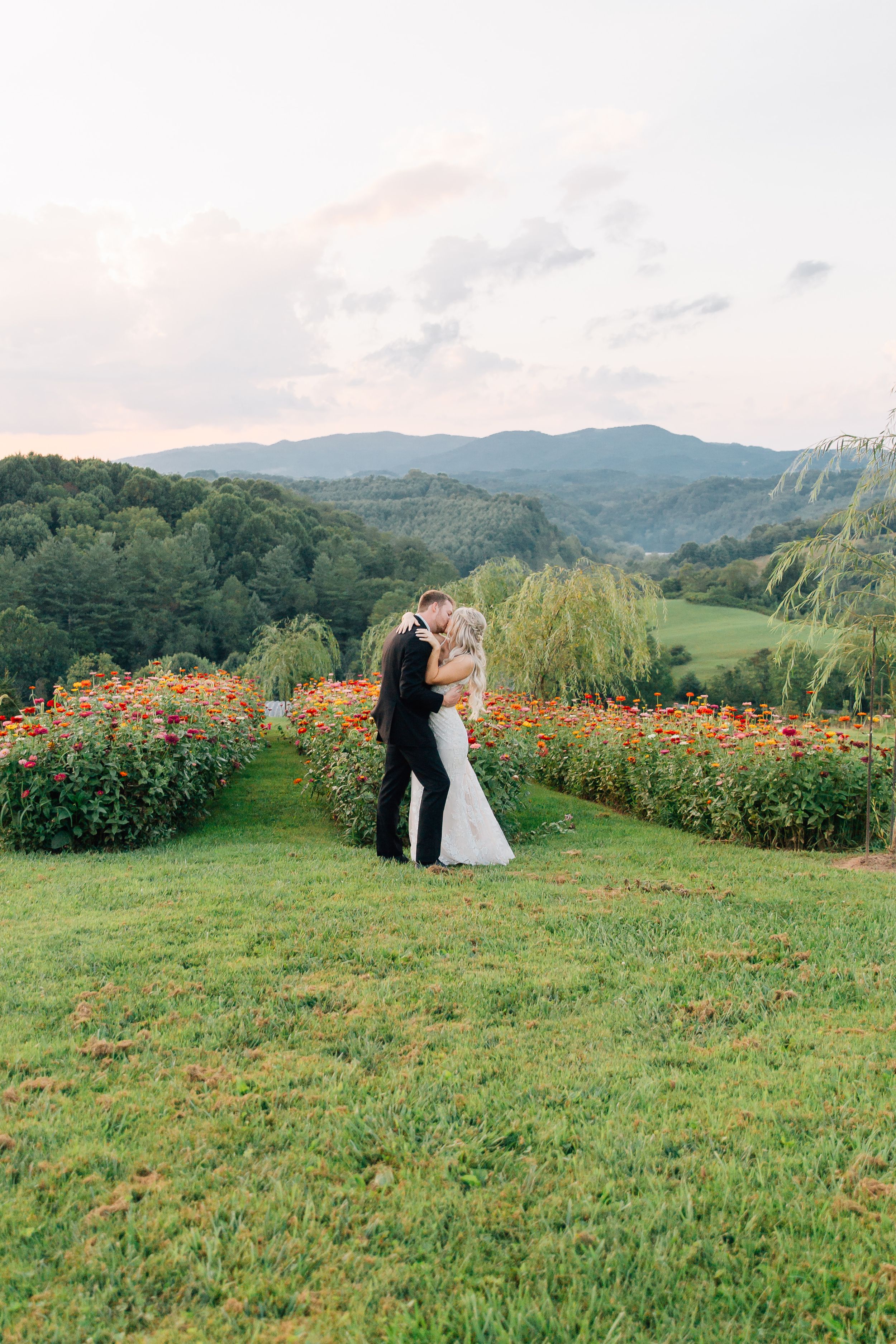 Wedding couple embraces in garden with mountain view and colorful flowers in background.