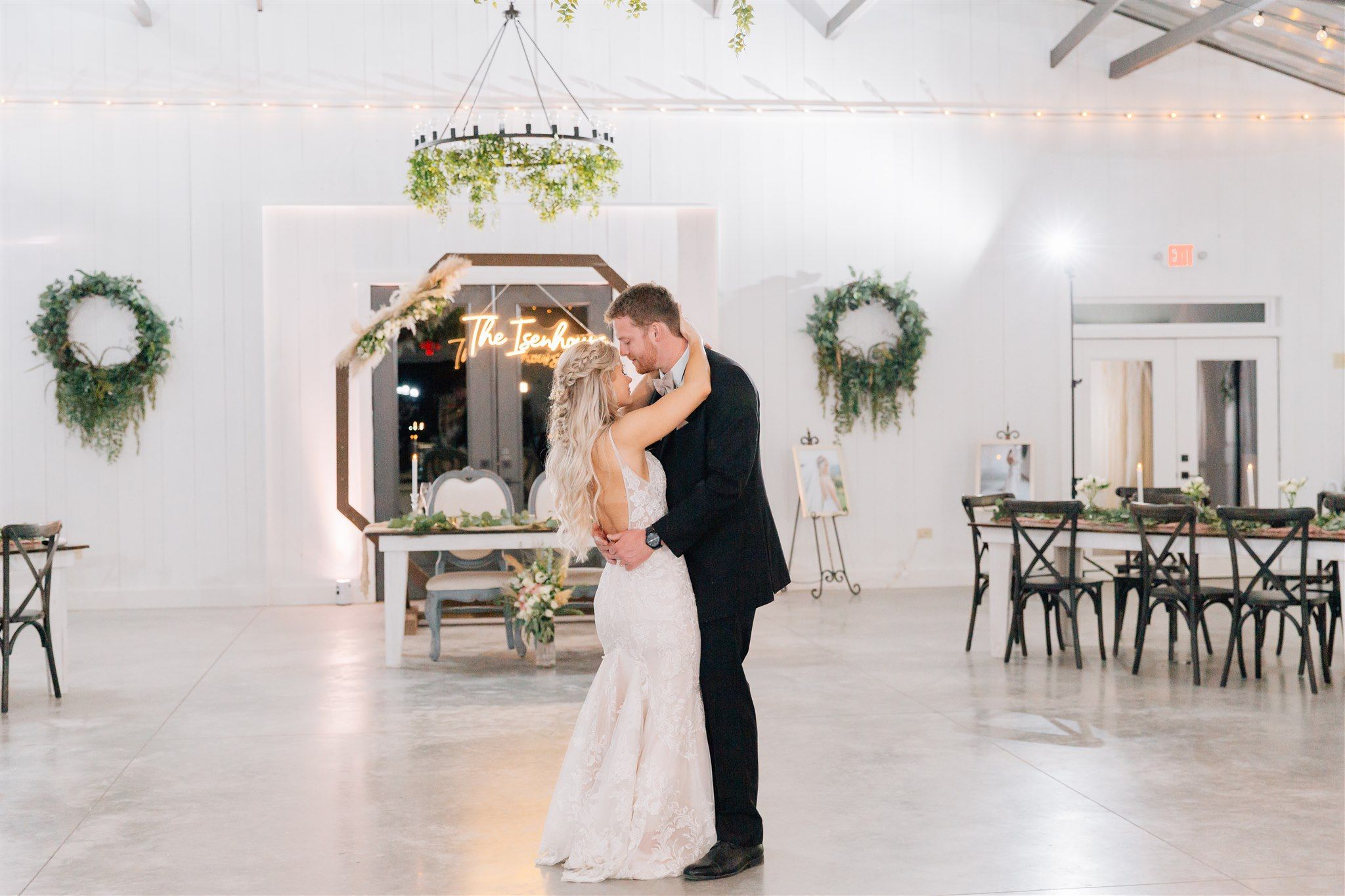 A couple shares their first dance in an elegant white reception venue decorated with greenery and chandeliers.