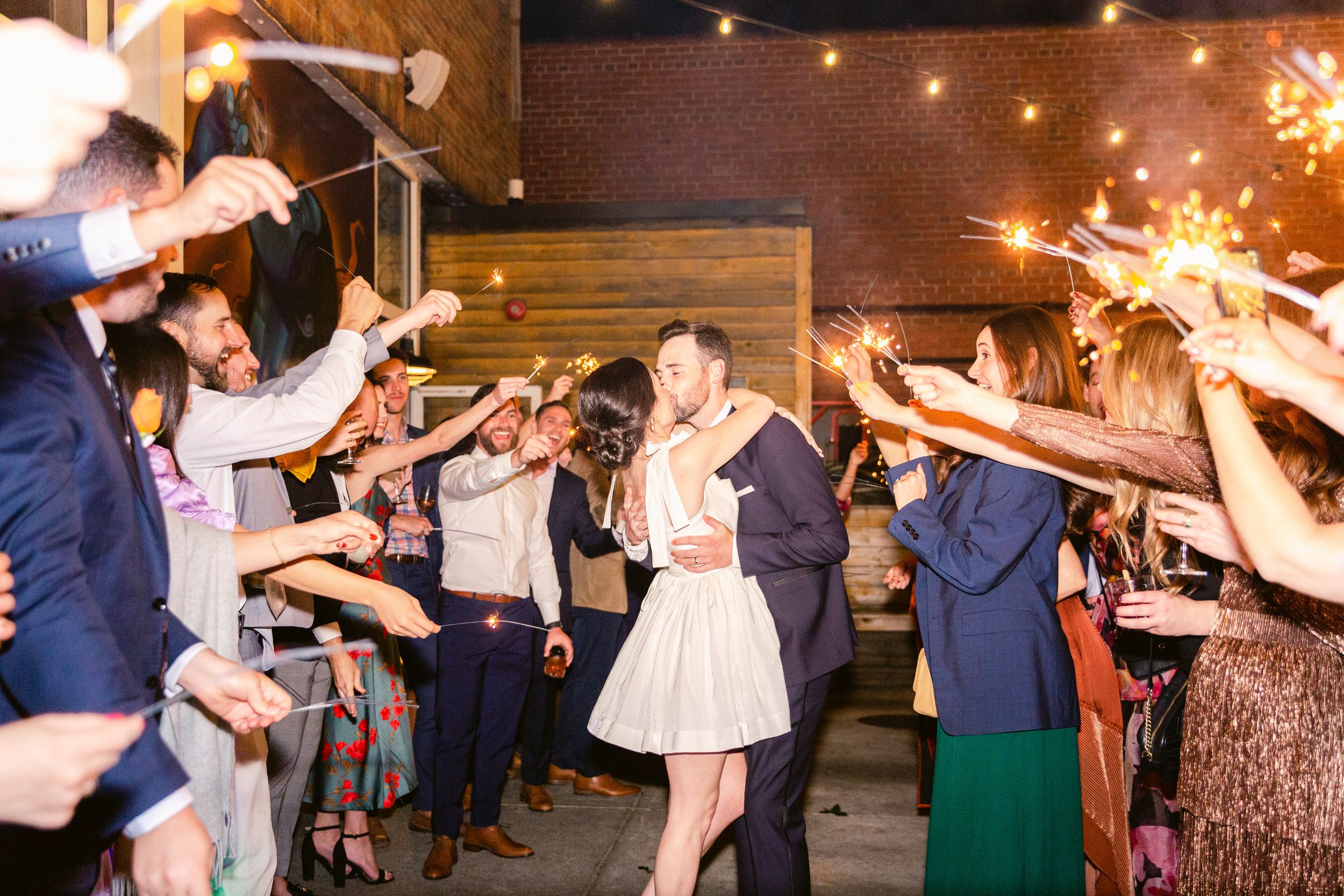 Wedding guests hold sparklers creating a magical nighttime sendoff tunnel outside a rustic venue.