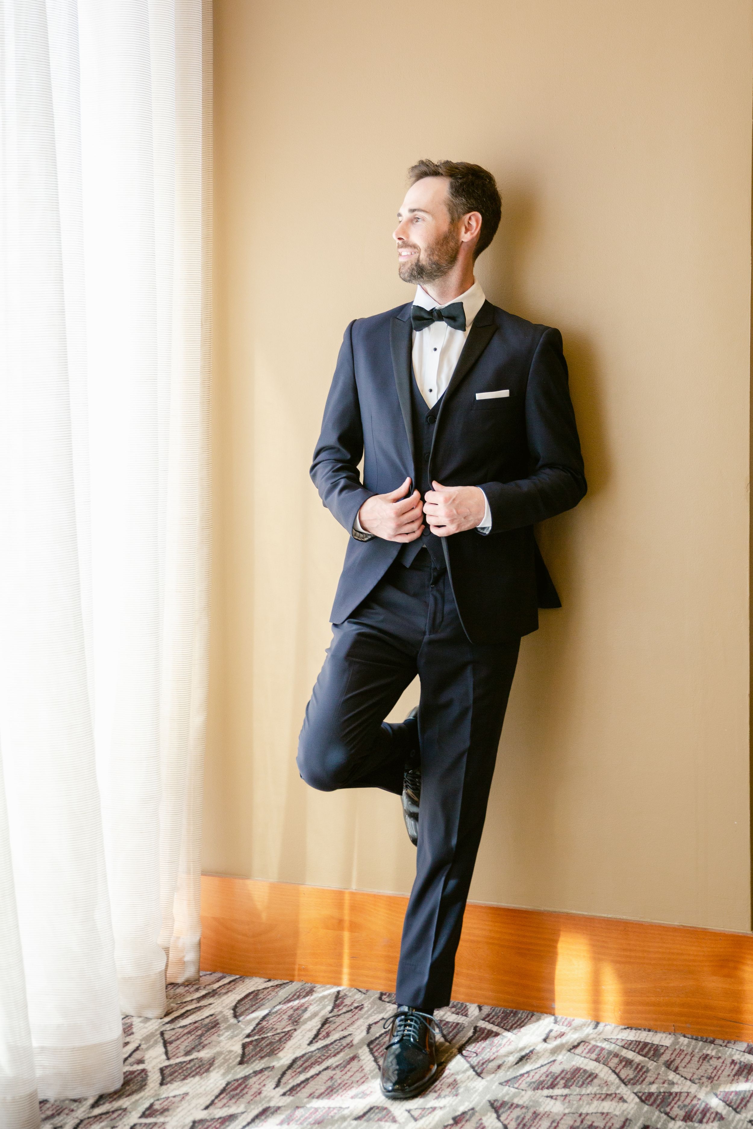 A groom in a classic black tuxedo and bow tie poses elegantly by a window while adjusting his jacket.