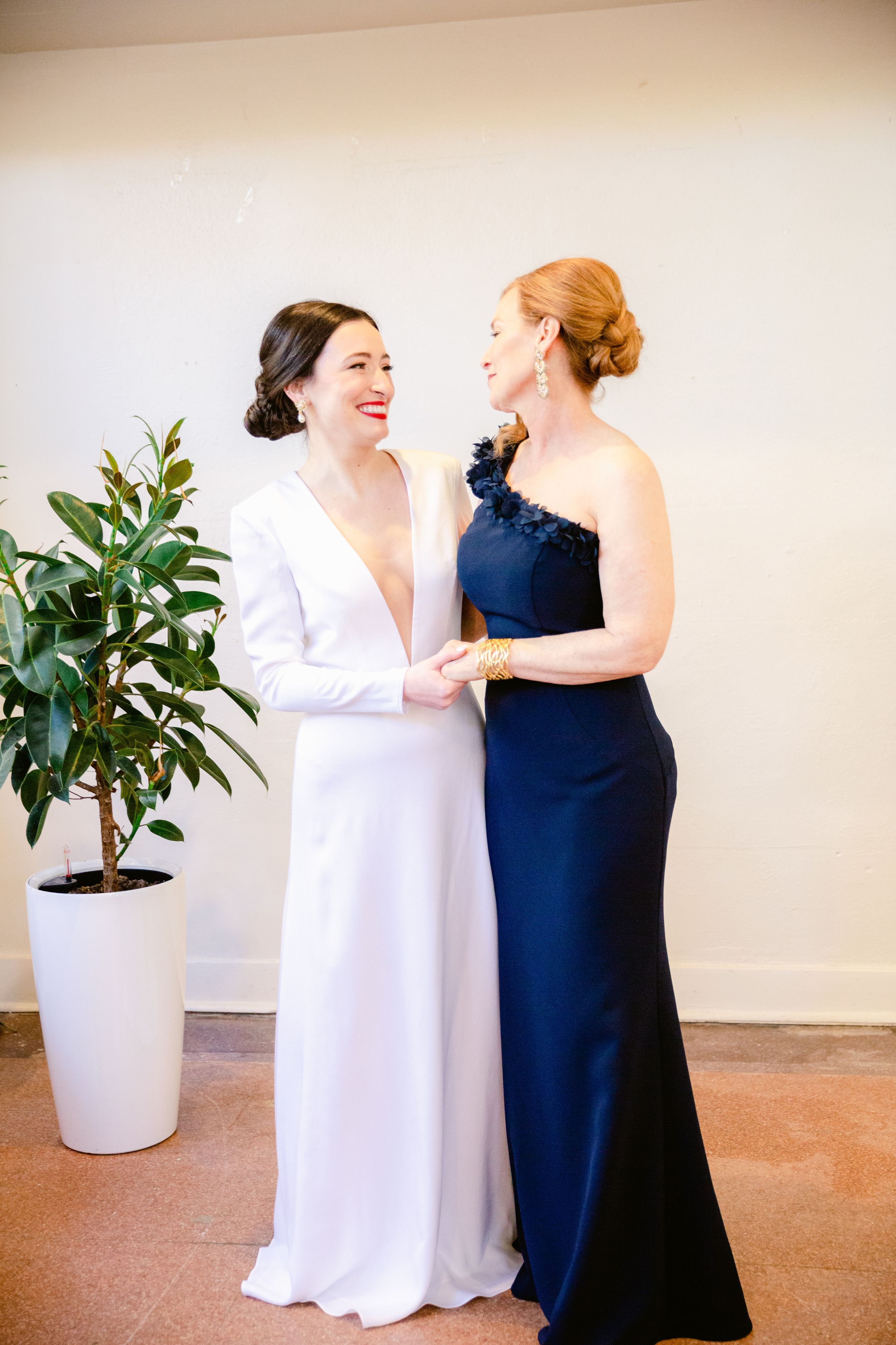 Two women in contrasting white and navy gowns share intimate moments in a minimalist setting with potted plants.