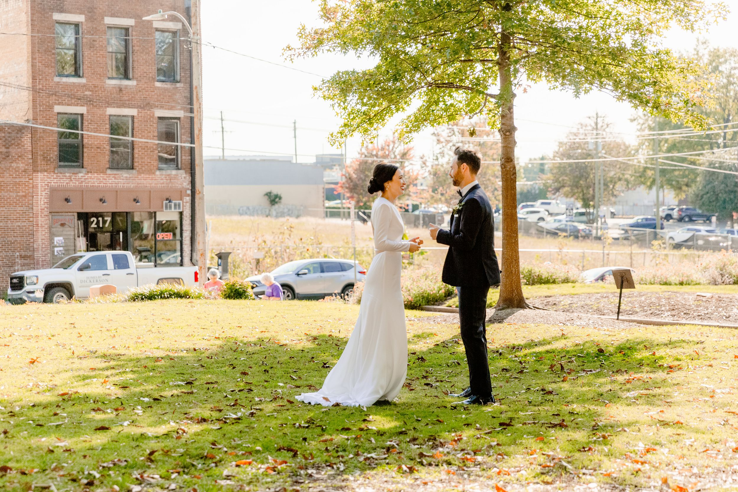 A couple in wedding attire stands on a grassy hill near brick buildings during golden hour.
