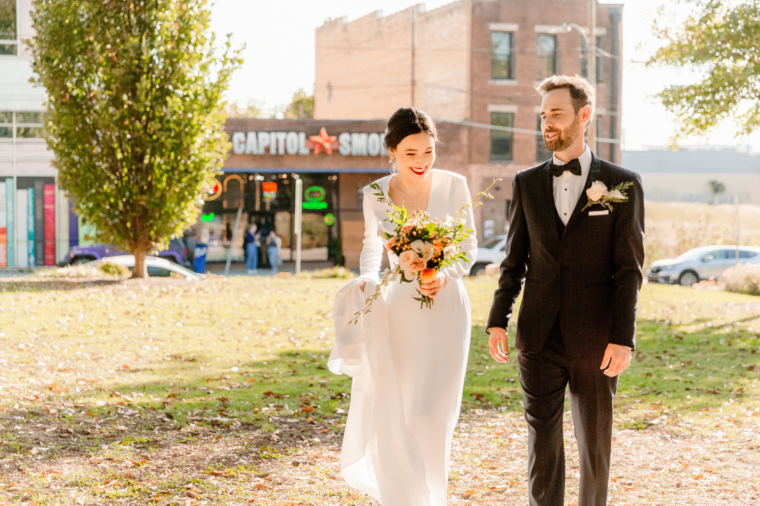 Bride and groom walk together through a park during autumn with fallen leaves scattered on the ground.