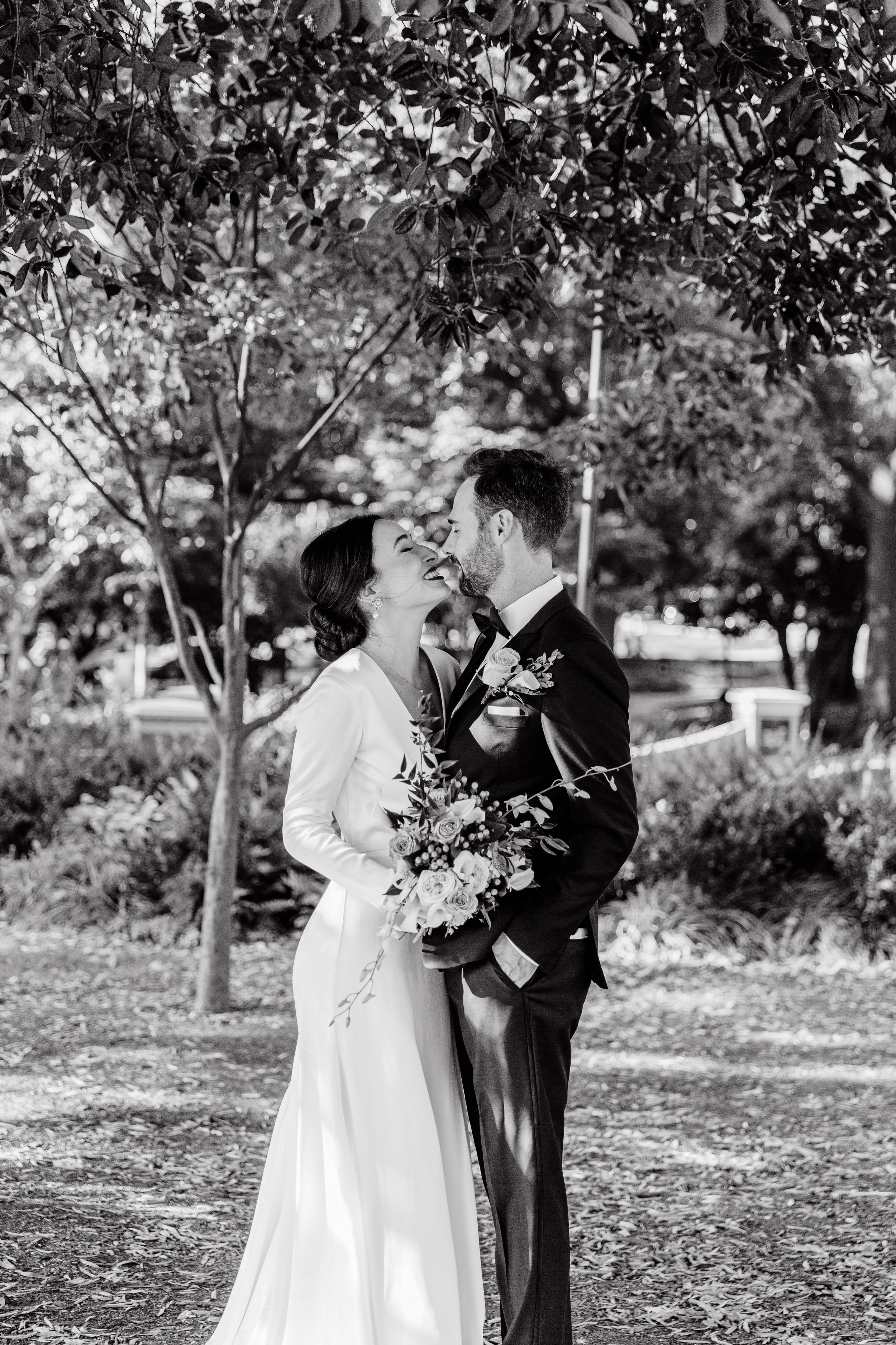 A romantic black and white wedding photo of a couple embracing under trees in a garden setting.