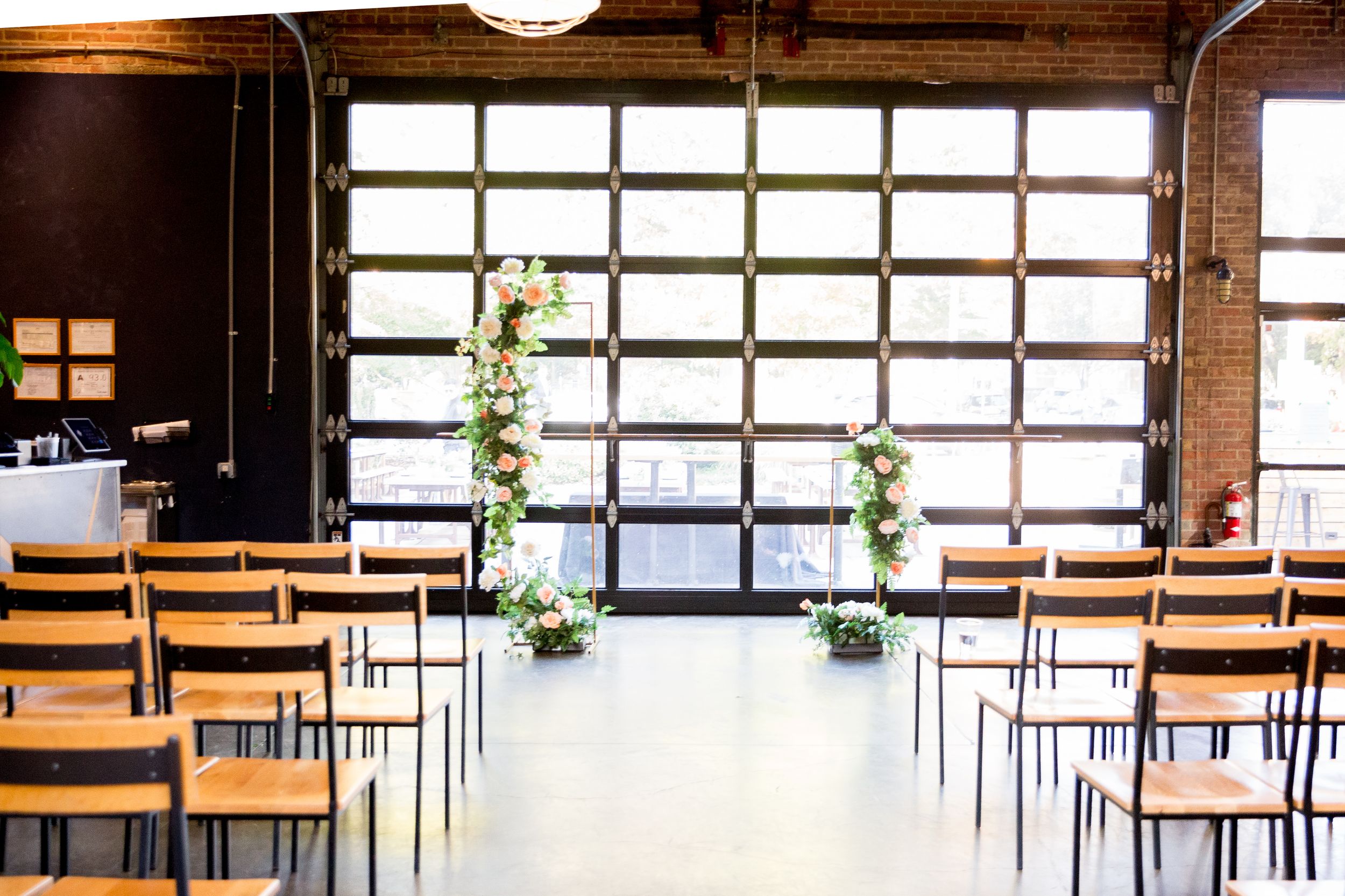 Modern industrial wedding ceremony space with wooden chairs and floral arrangements along the aisle near a glass garage door.