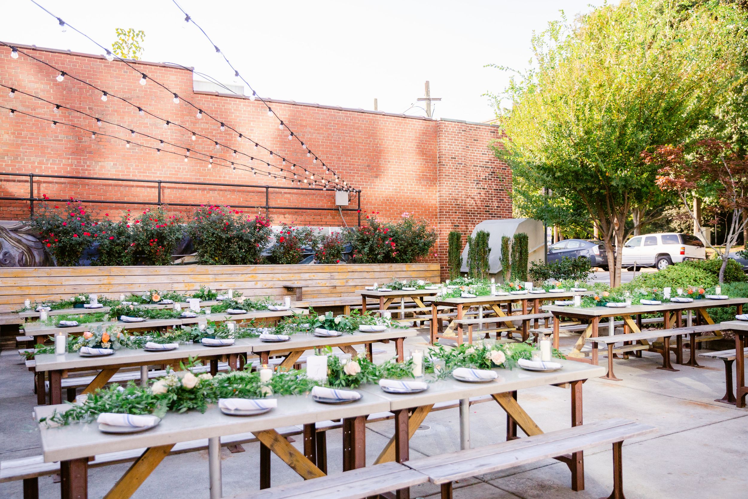 A panoramic view of outdoor picnic tables and benches arranged in rows next to a brick building and garden area.