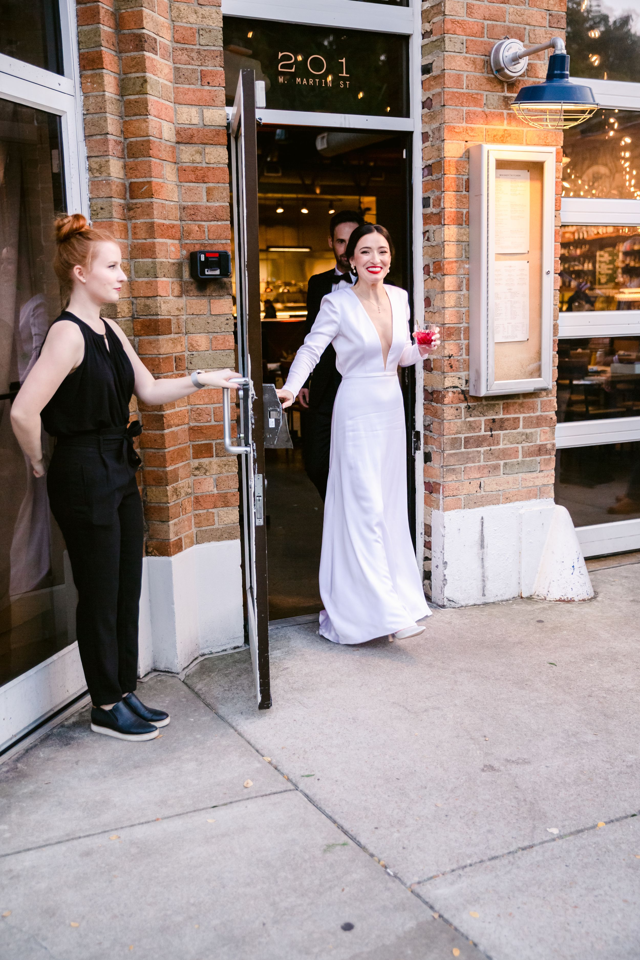 Two people in formal attire playfully interact outside a brick storefront entrance.