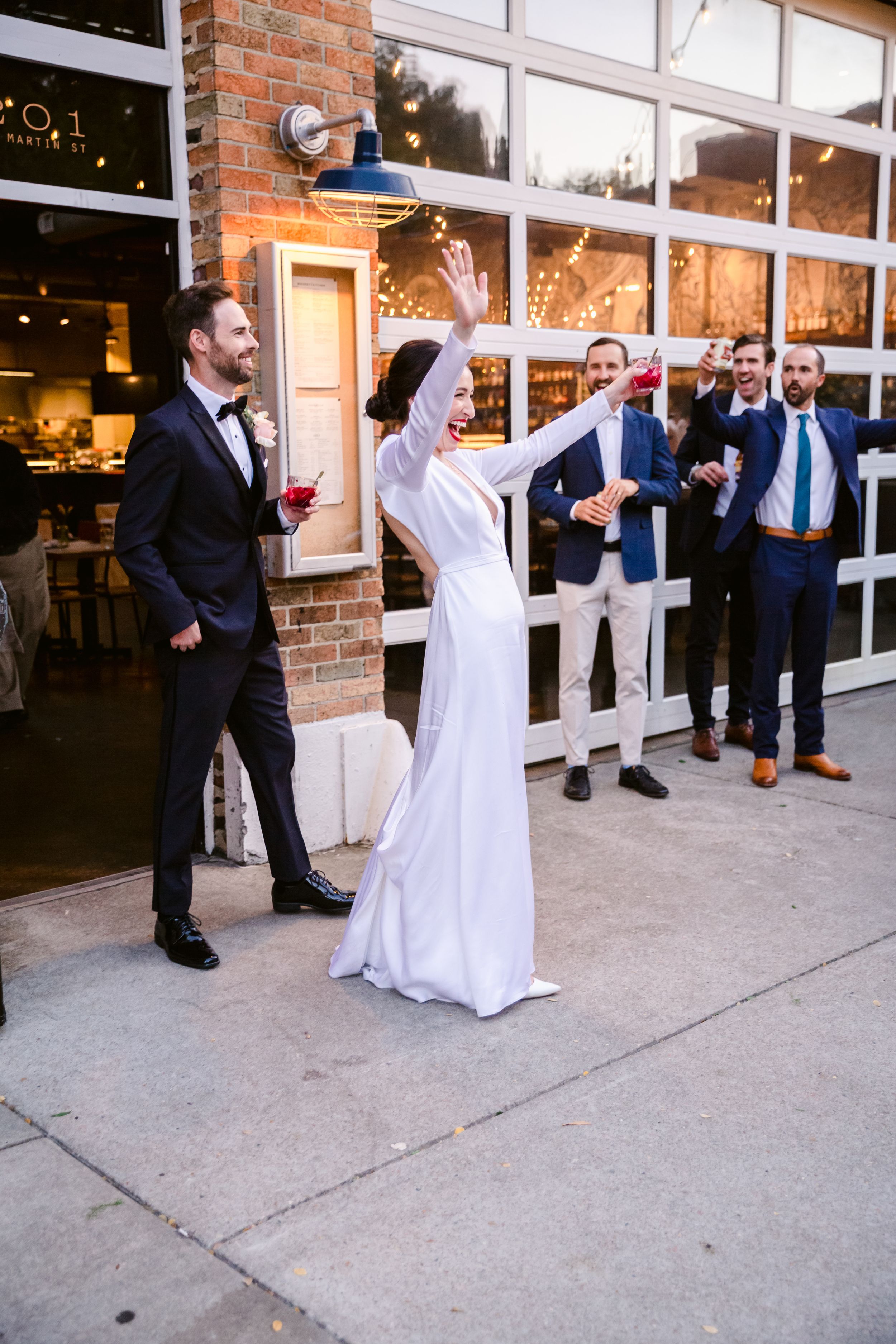 Wedding celebration outside a brick venue with guests cheering as the bride raises her arms in joy.