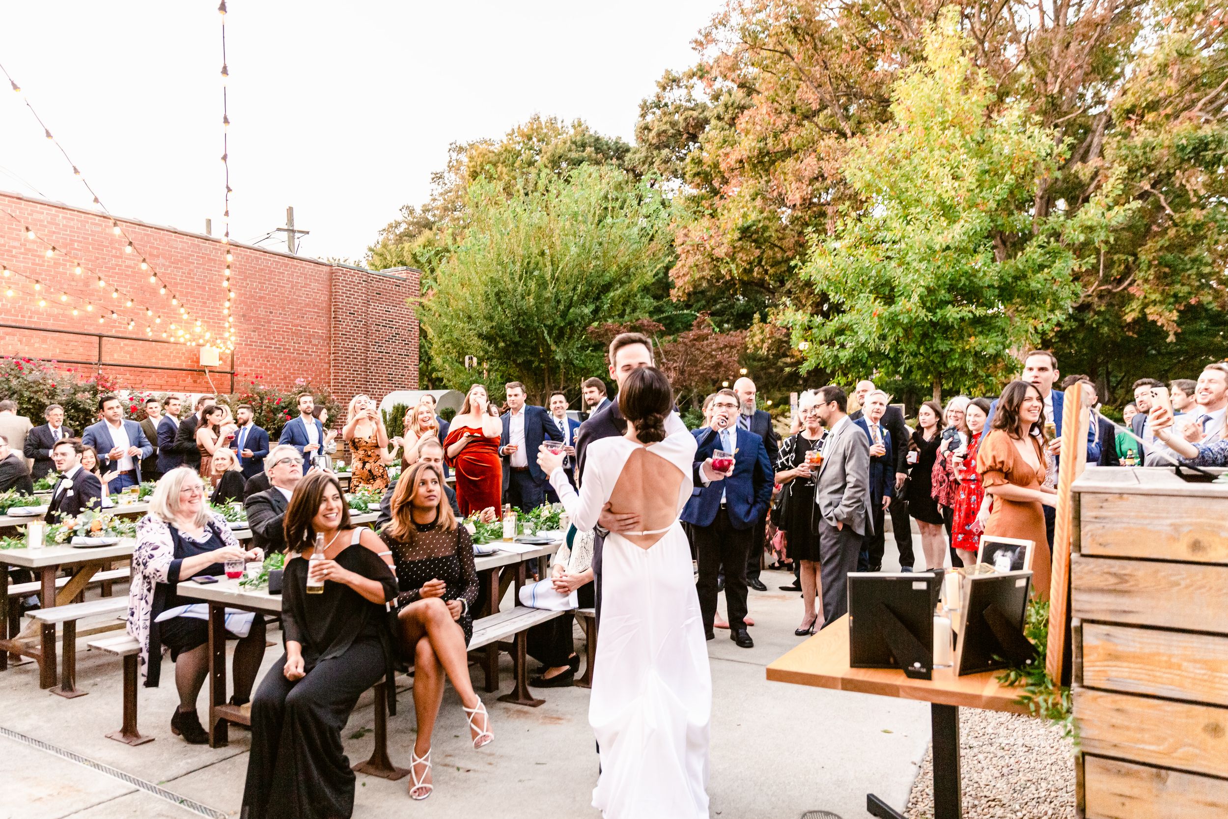A crowd gathers outside a brick building with trees and landscaping during a daytime event.