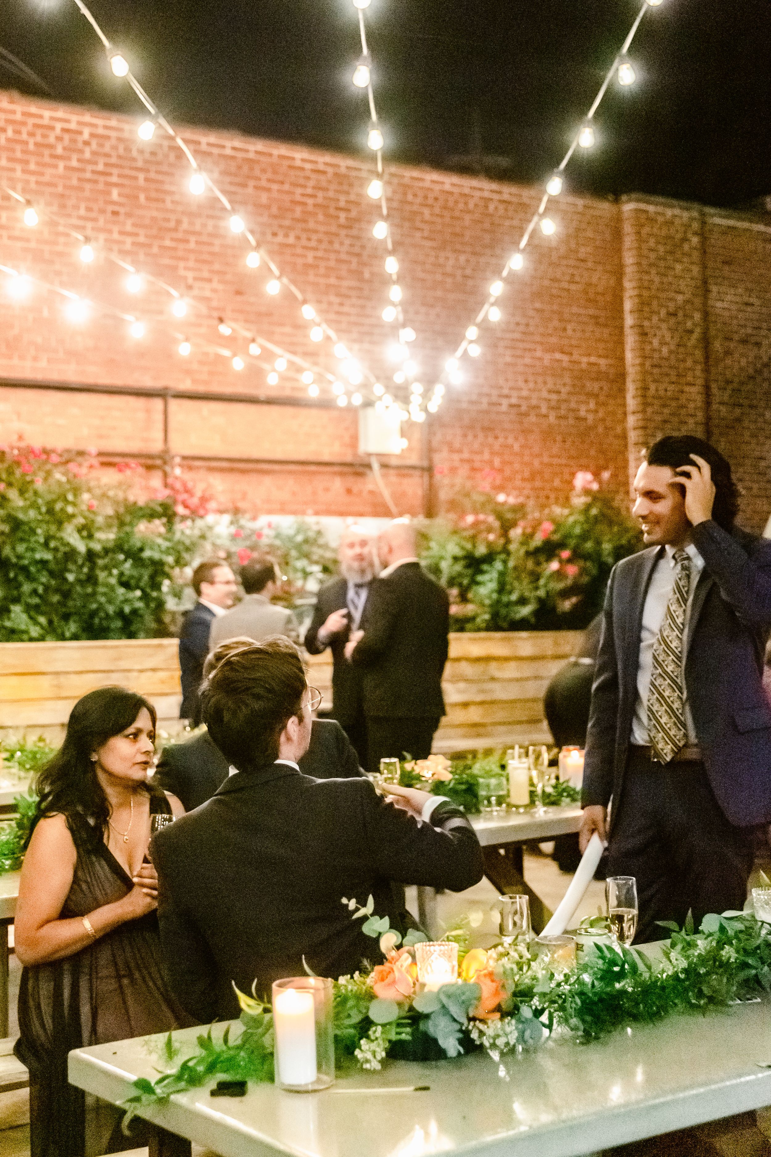 People gather at an outdoor evening event under string lights with candles and greenery decorating long tables.