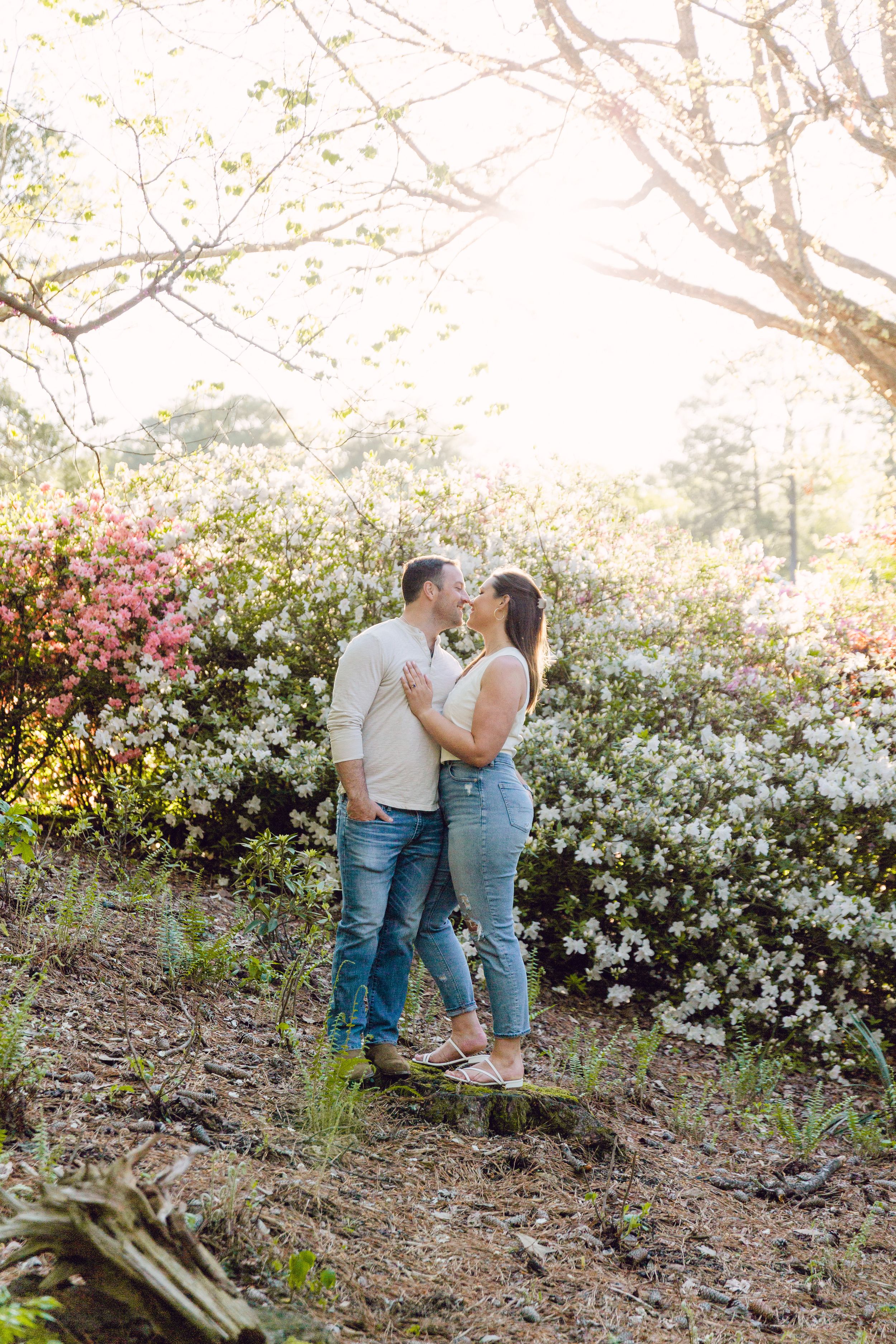 A couple embraces among blooming spring flowers and trees in a sunlit garden setting.