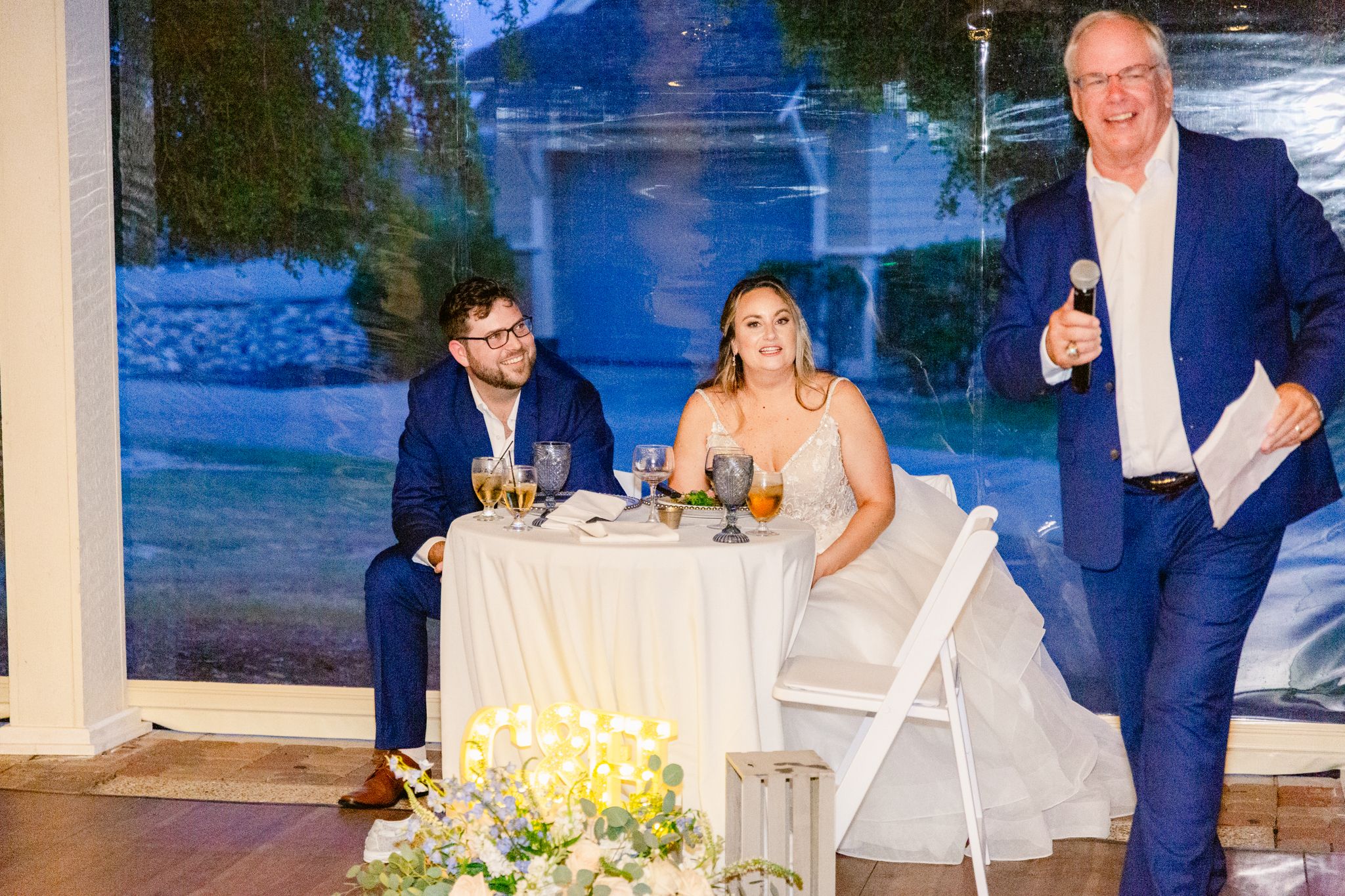 Wedding reception guests seated at outdoor tables with blue lighting and floral centerpieces at sunset.
