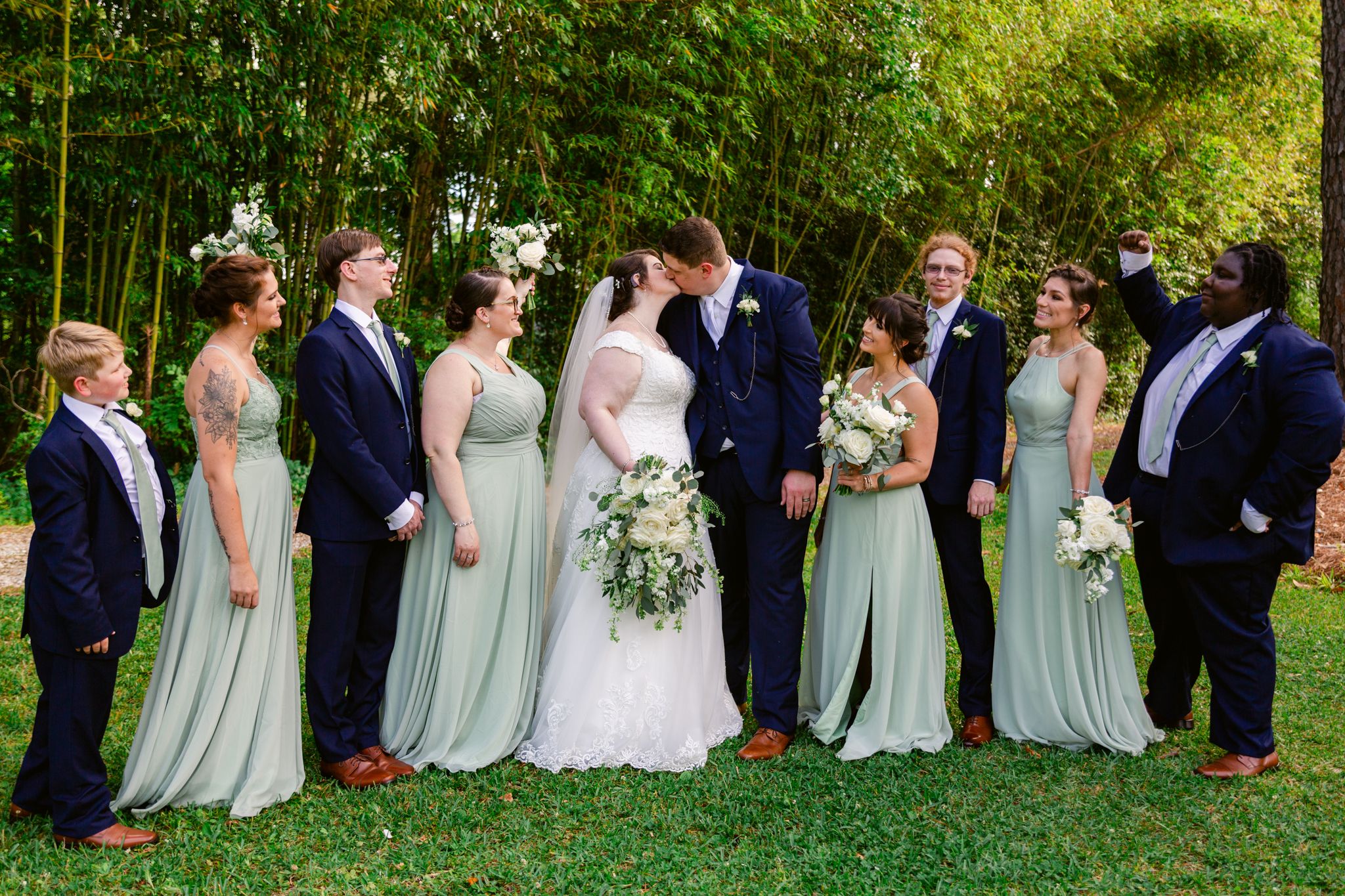 A wedding party in navy suits and sage green bridesmaid dresses poses outdoors in a line against lush greenery.