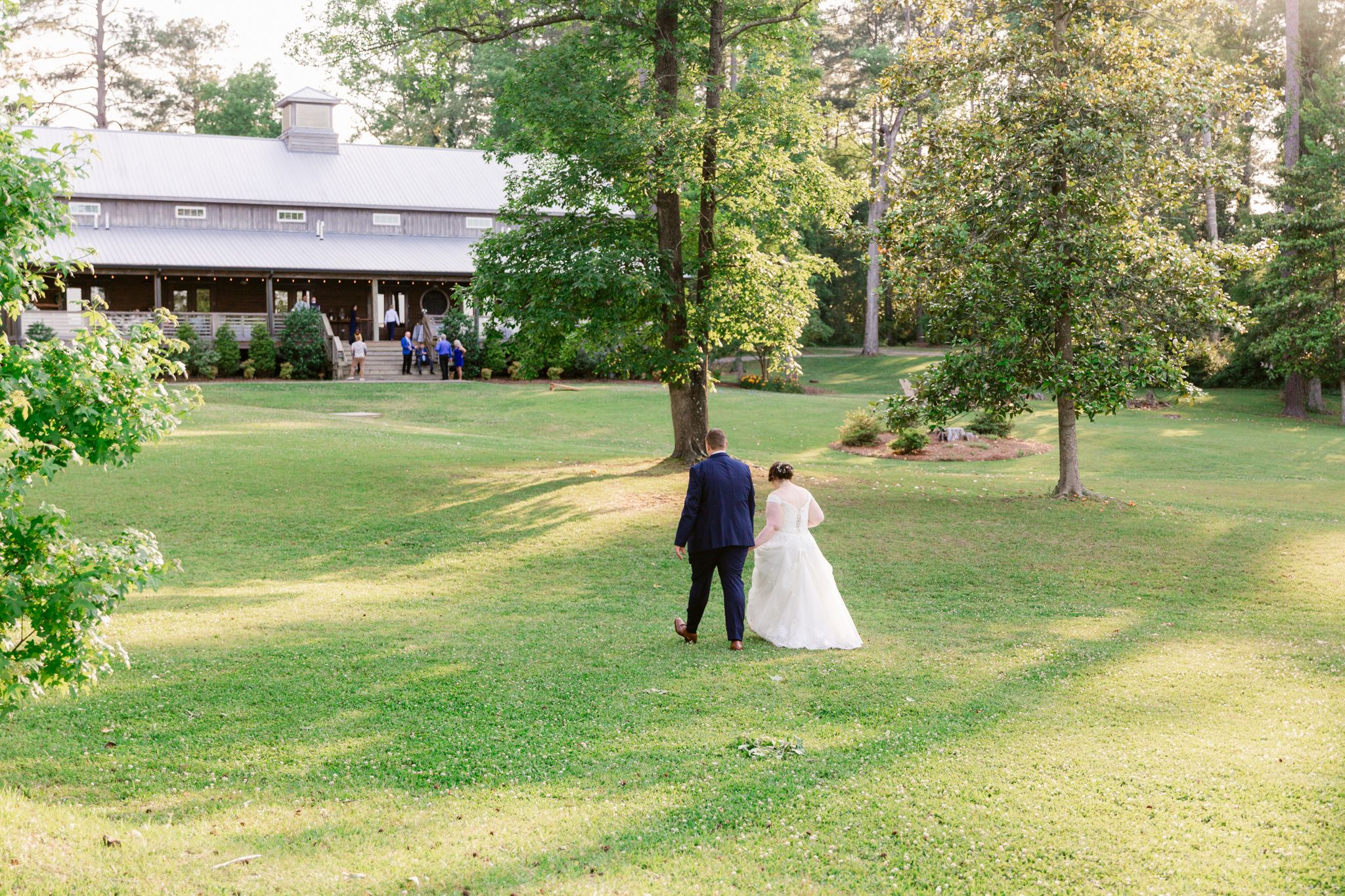 A couple stands together on a lush green lawn near a rustic wooden lodge surrounded by trees.