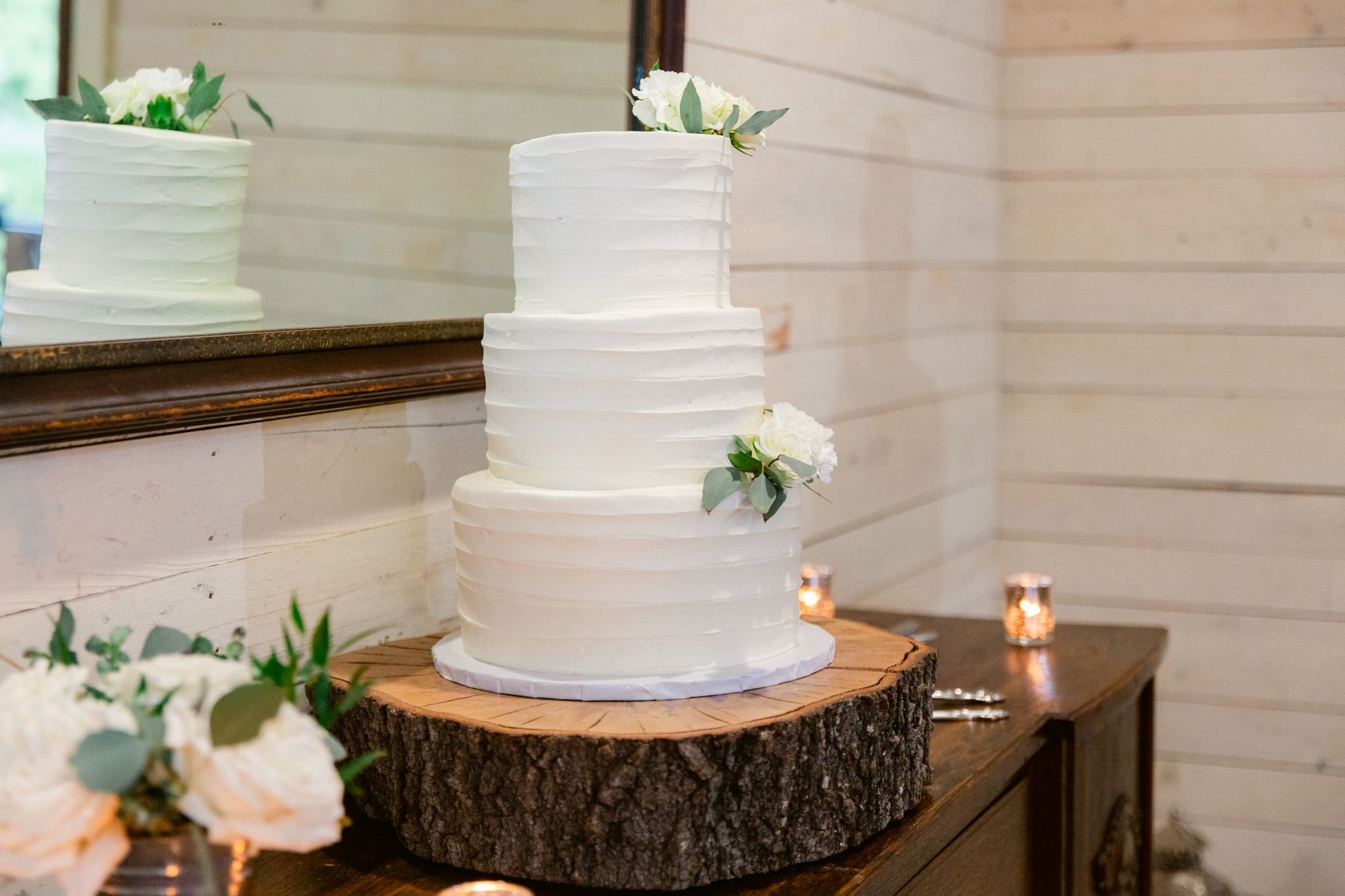 Elegant white tiered wedding cake displayed on rustic wood slice with delicate floral accents and candlelight.