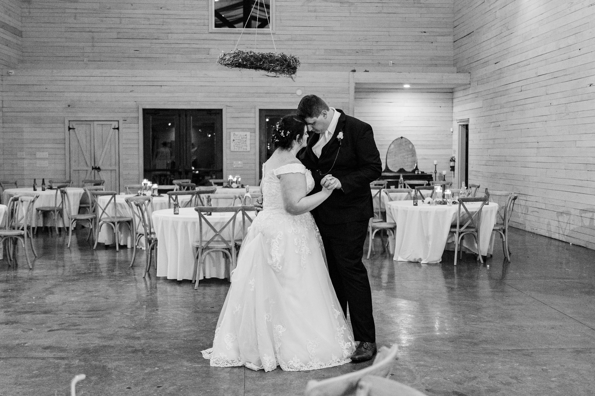 A couple shares their first dance in a rustic white-walled reception venue with round tables and cross-back chairs.