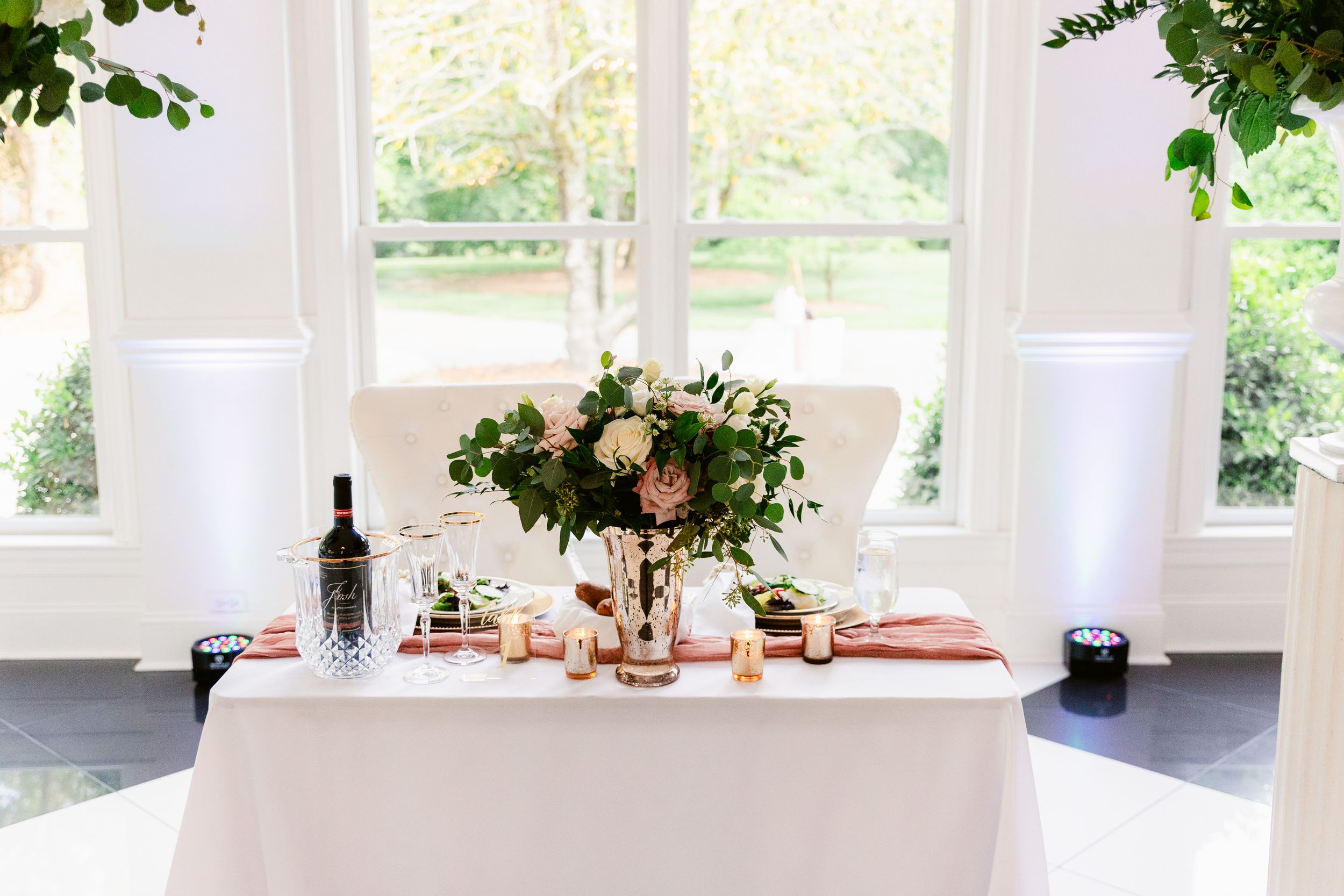 Elegant white dining room with large windows, natural light, and green foliage accents creating an airy atmosphere.