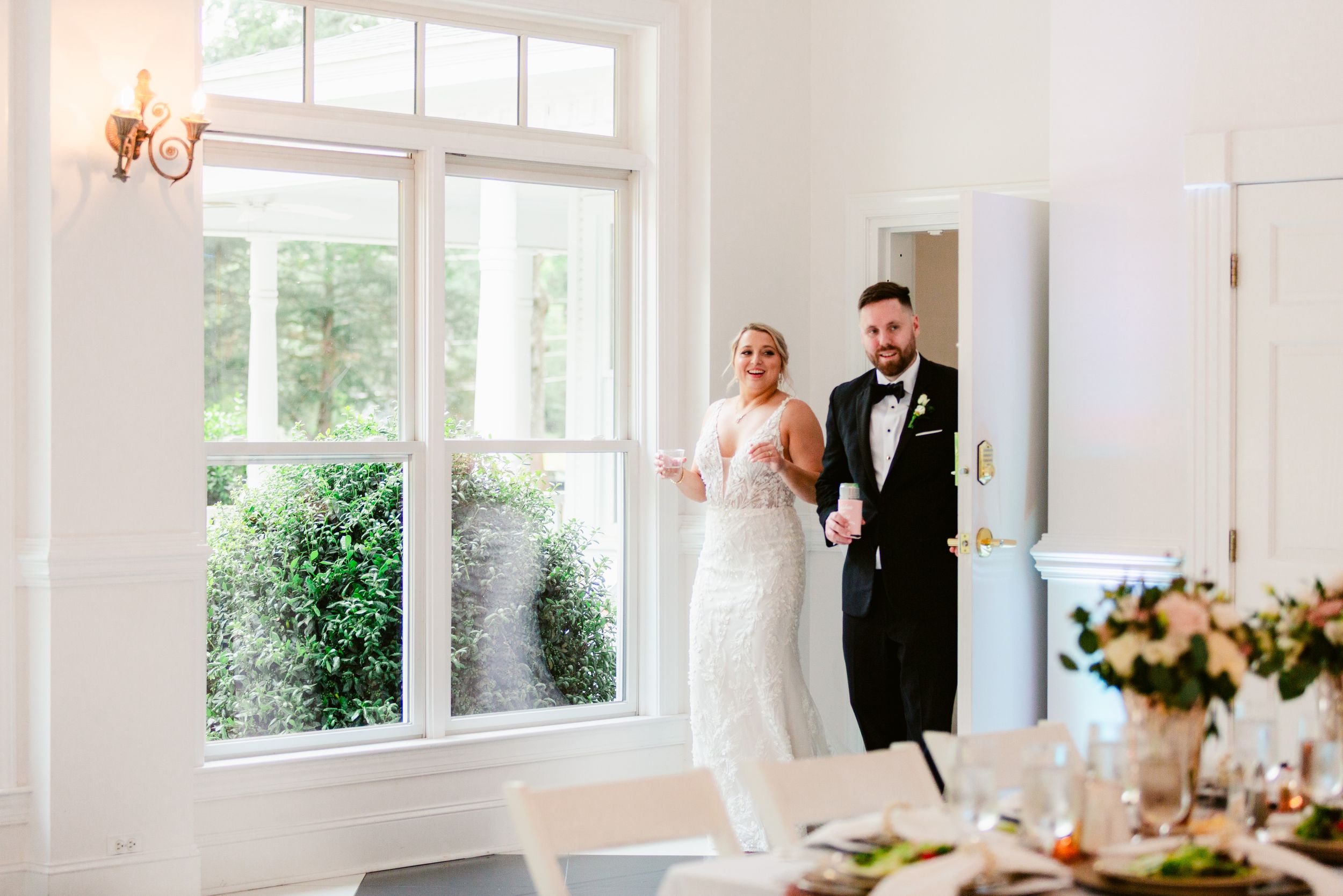 Elegant white wedding reception venue with large windows, floral centerpieces, and natural light streaming in.
