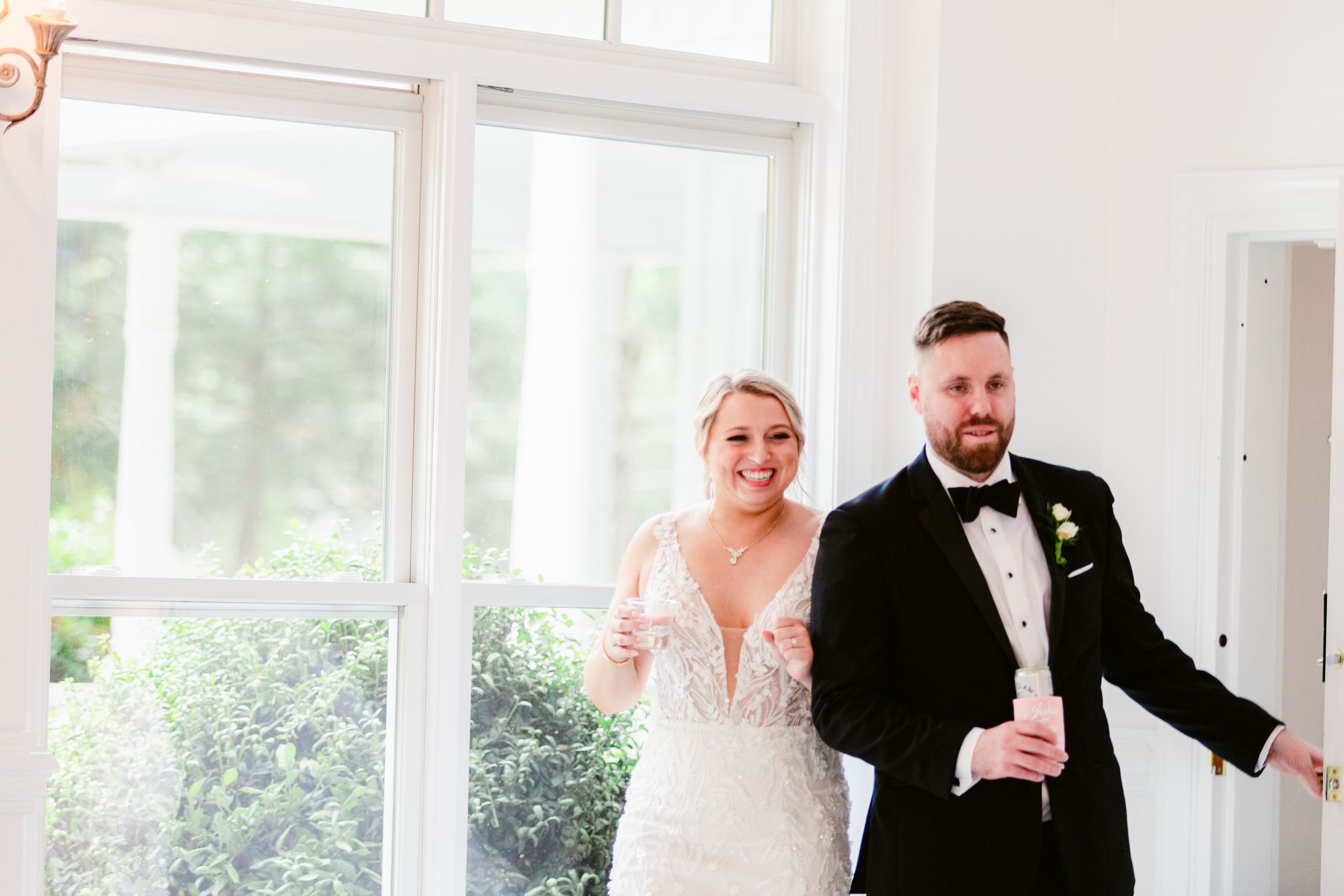 A wedding party stands together in a bright, sunlit room with white decor and large windows.