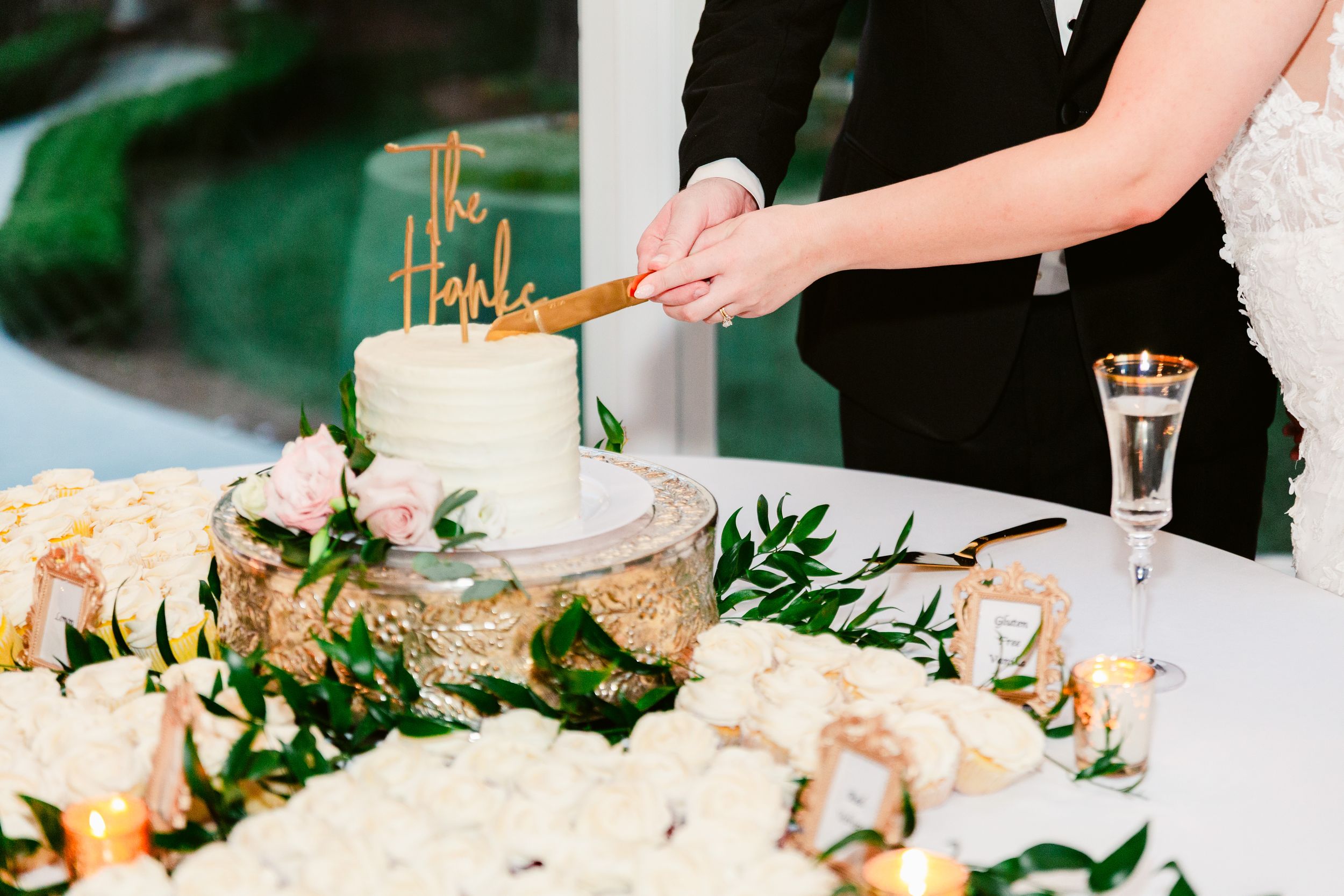 Hands cut into an elegant white wedding cake adorned with fresh flowers and greenery at a formal reception.