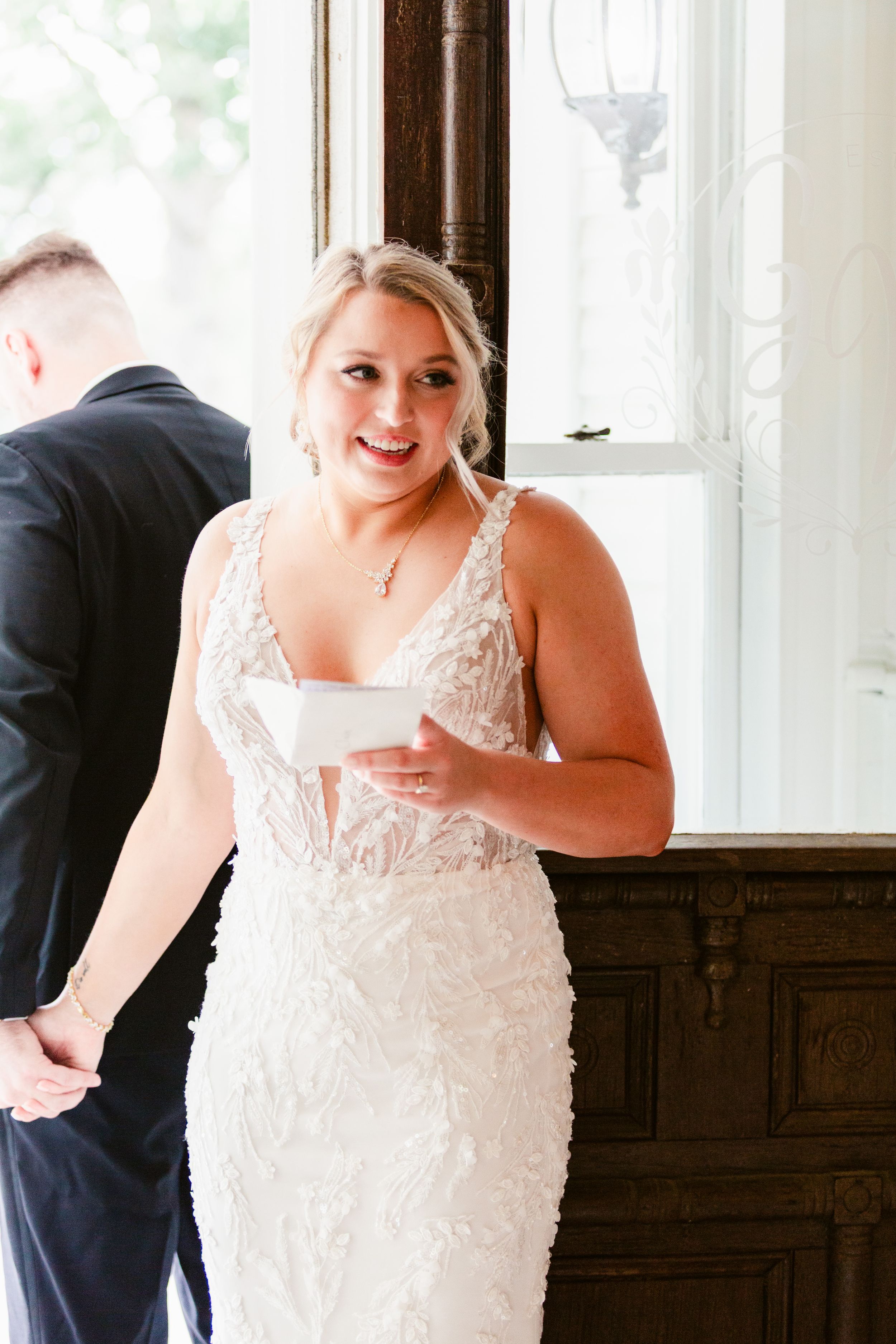 Bride in an elegant white lace wedding gown with delicate straps holds a glass during a reception.