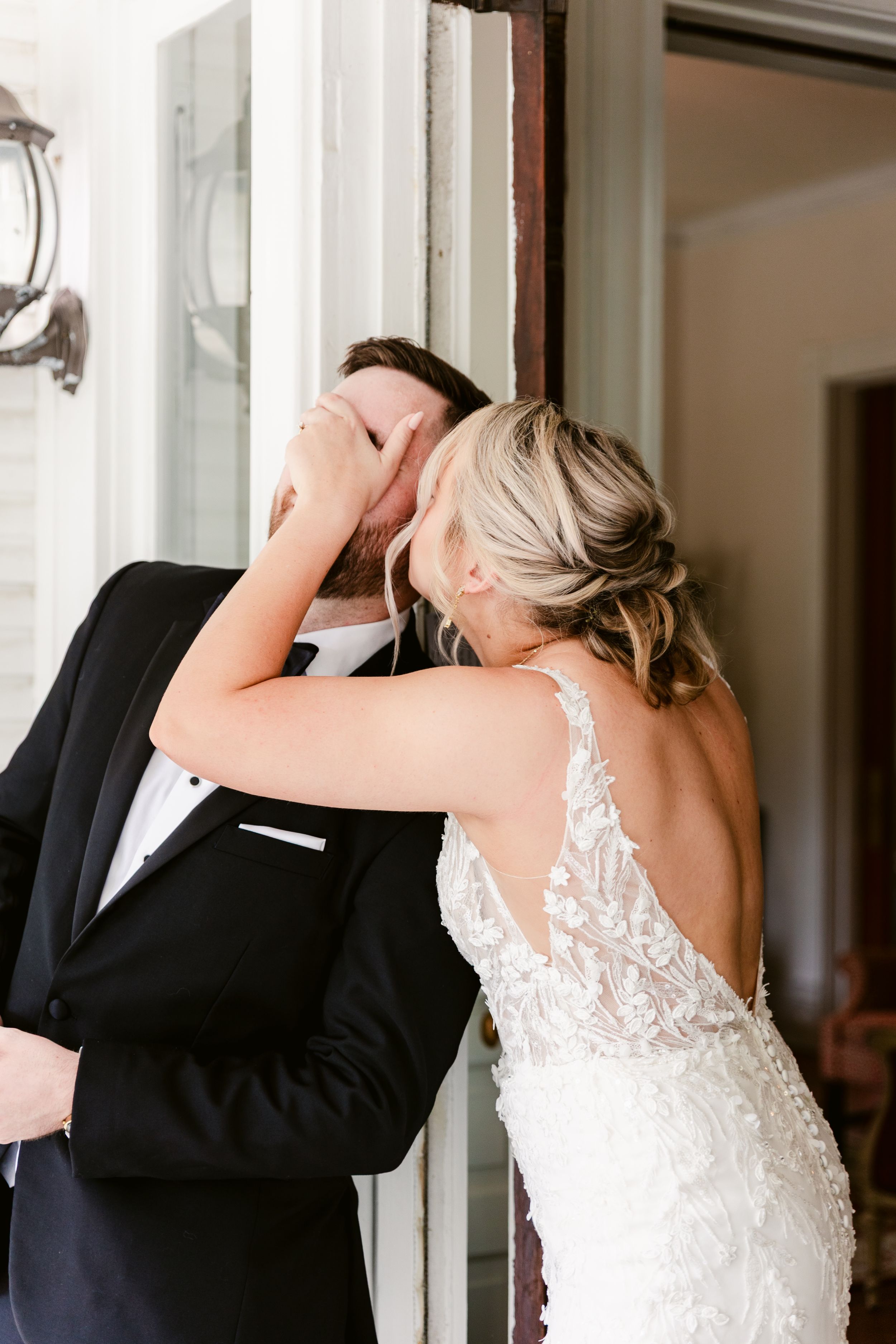 Bride in a fitted lace gown shares a romantic first look moment with groom near a doorway.