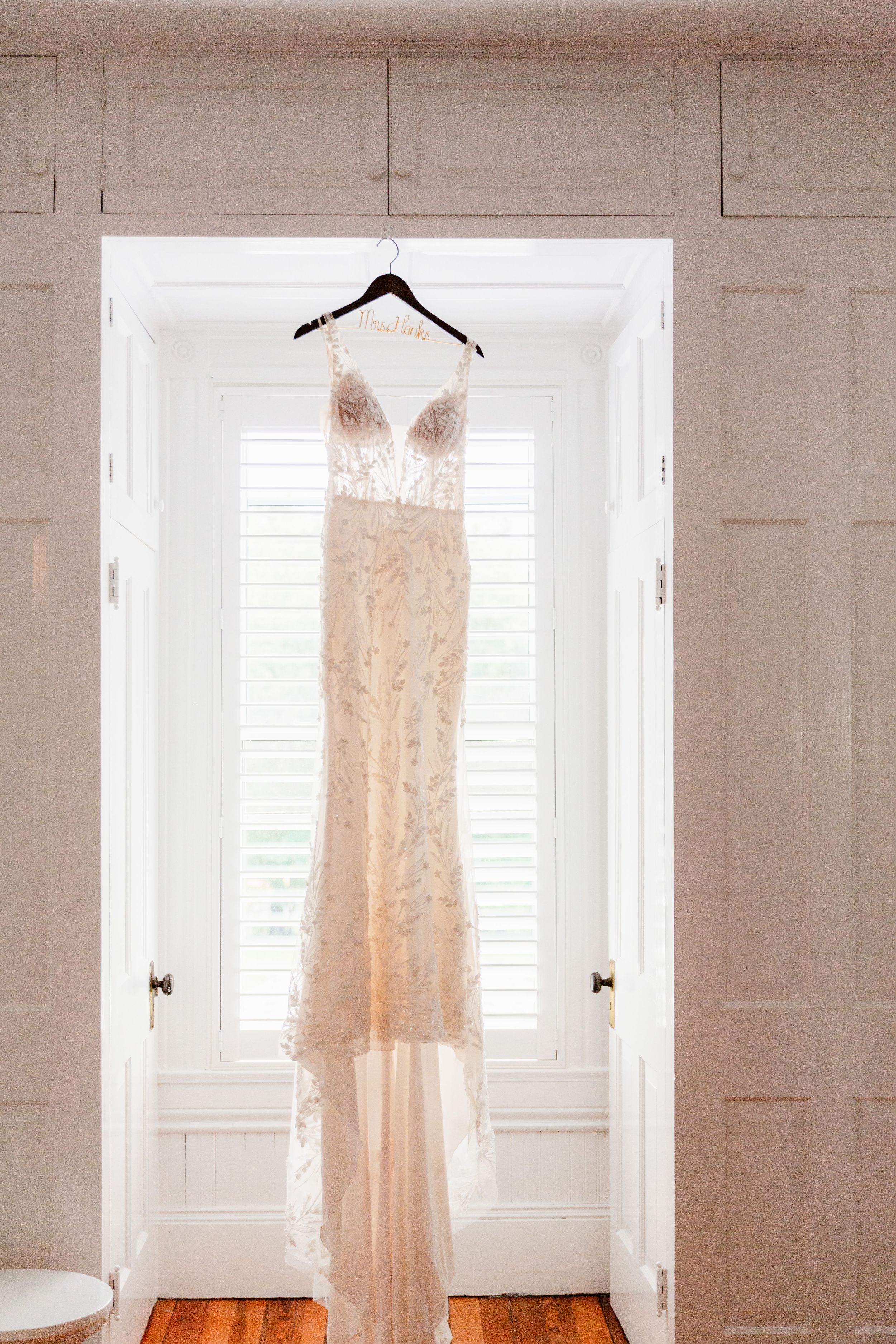 Elegant lace wedding dress hanging in white room with natural light streaming through windows.