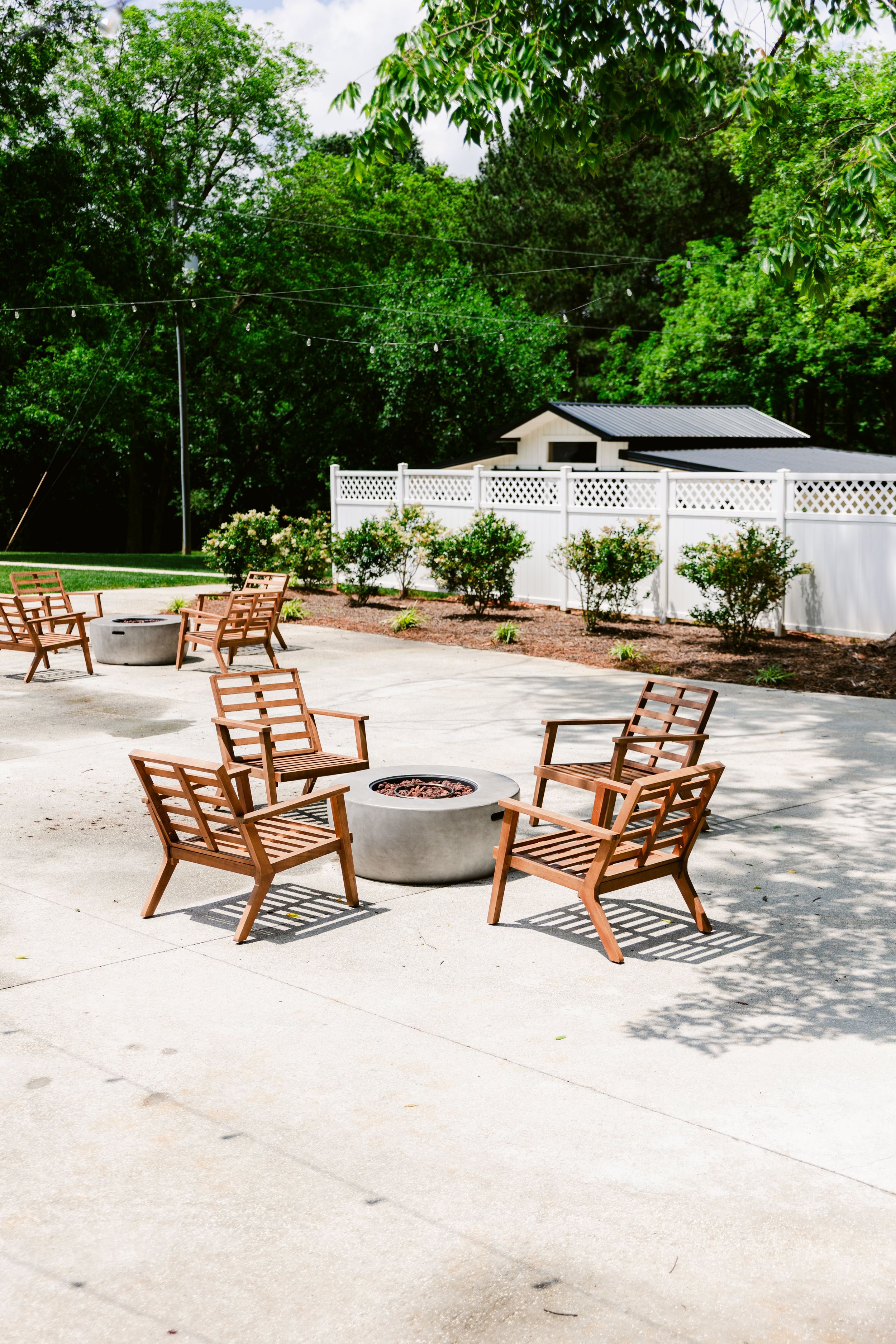 Wooden lounge chairs arranged around a fire pit on a concrete patio surrounded by greenery.