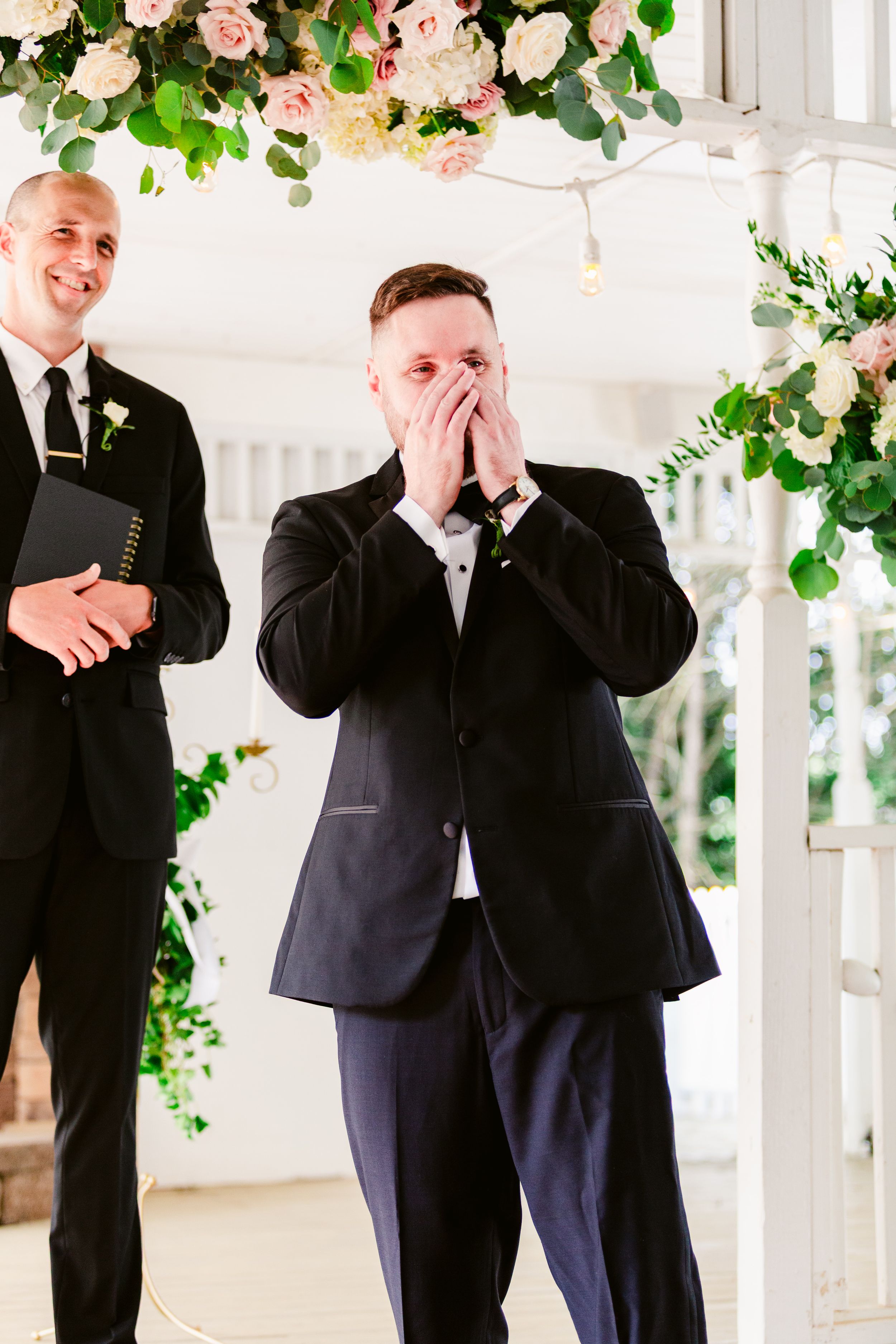 Two people in formal black suits sharing a joyful moment under floral decorations.