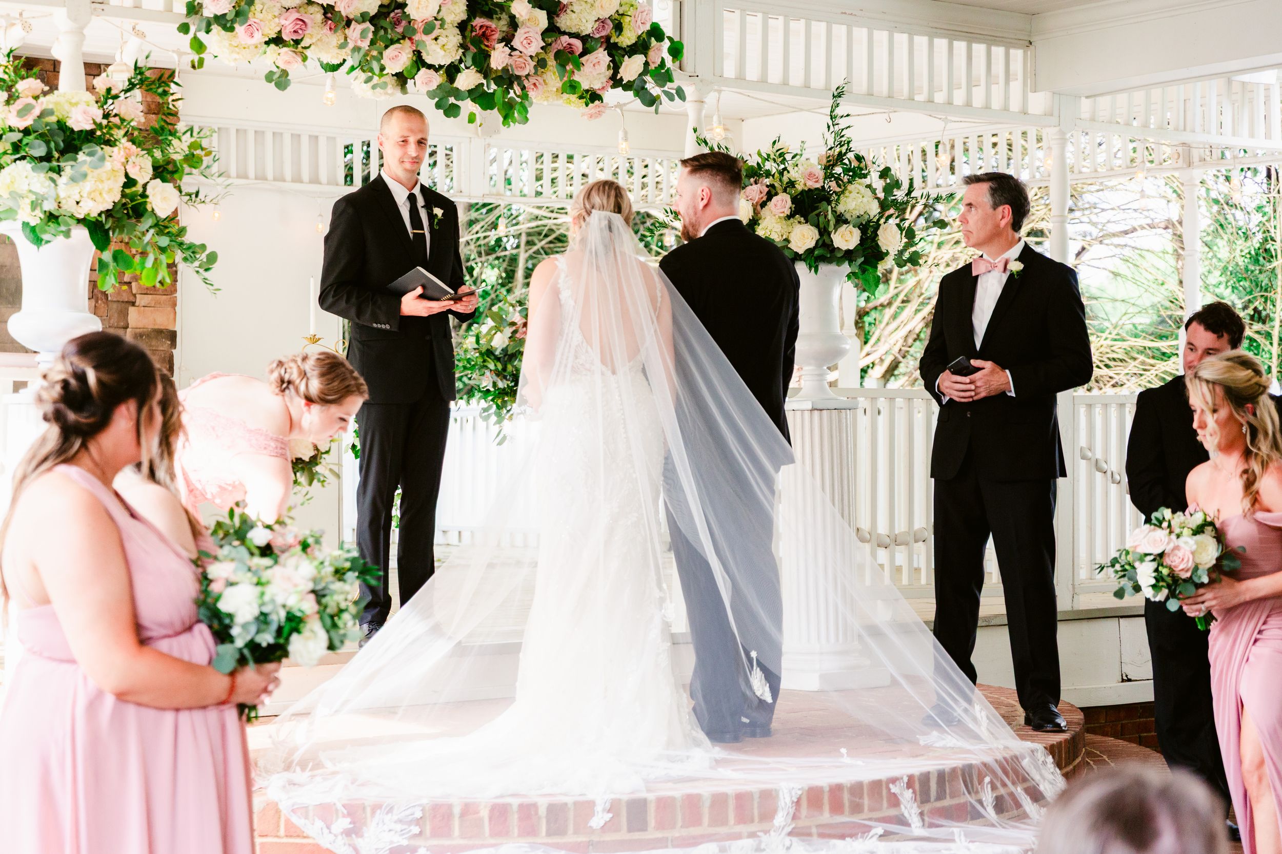Wedding party gathers on a white porch decorated with pink and white floral arrangements for formal photographs.