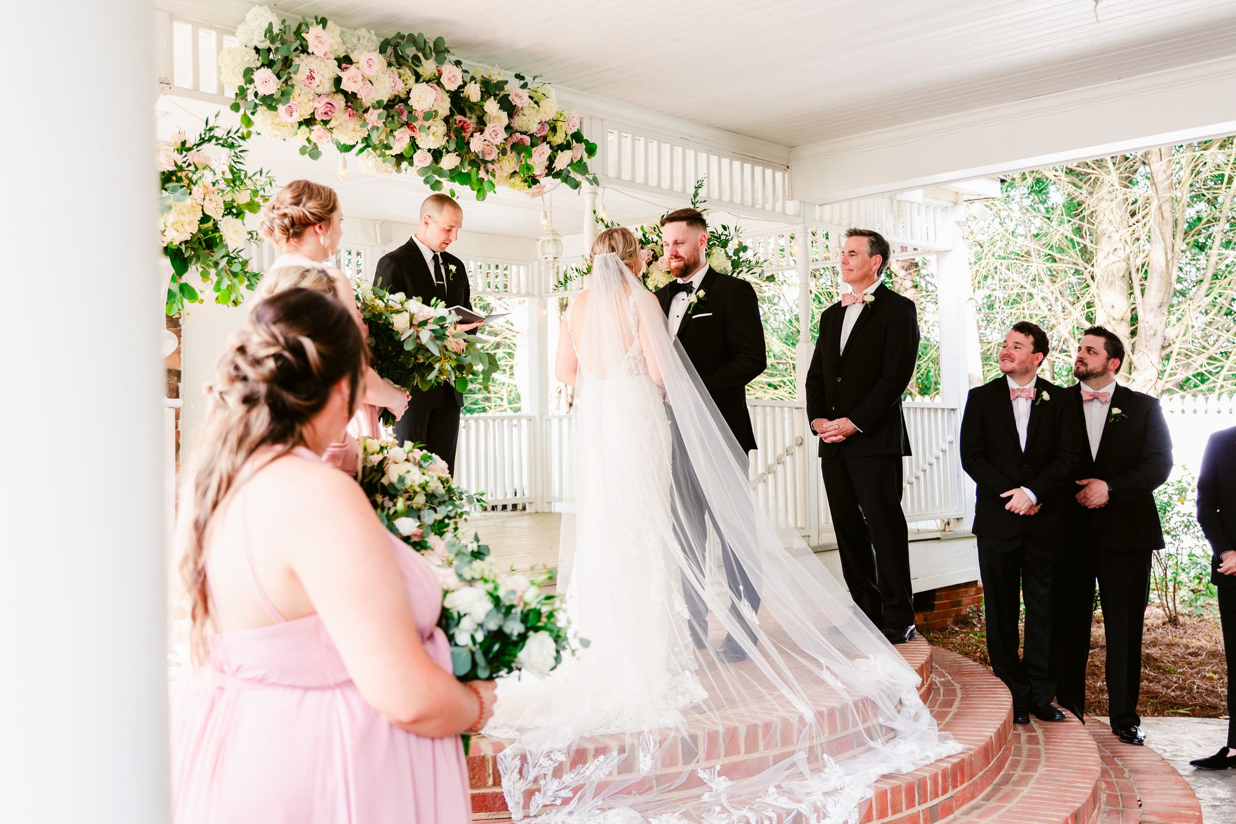 Wedding ceremony under a white covered porch with floral arch and pink bridesmaid dresses.