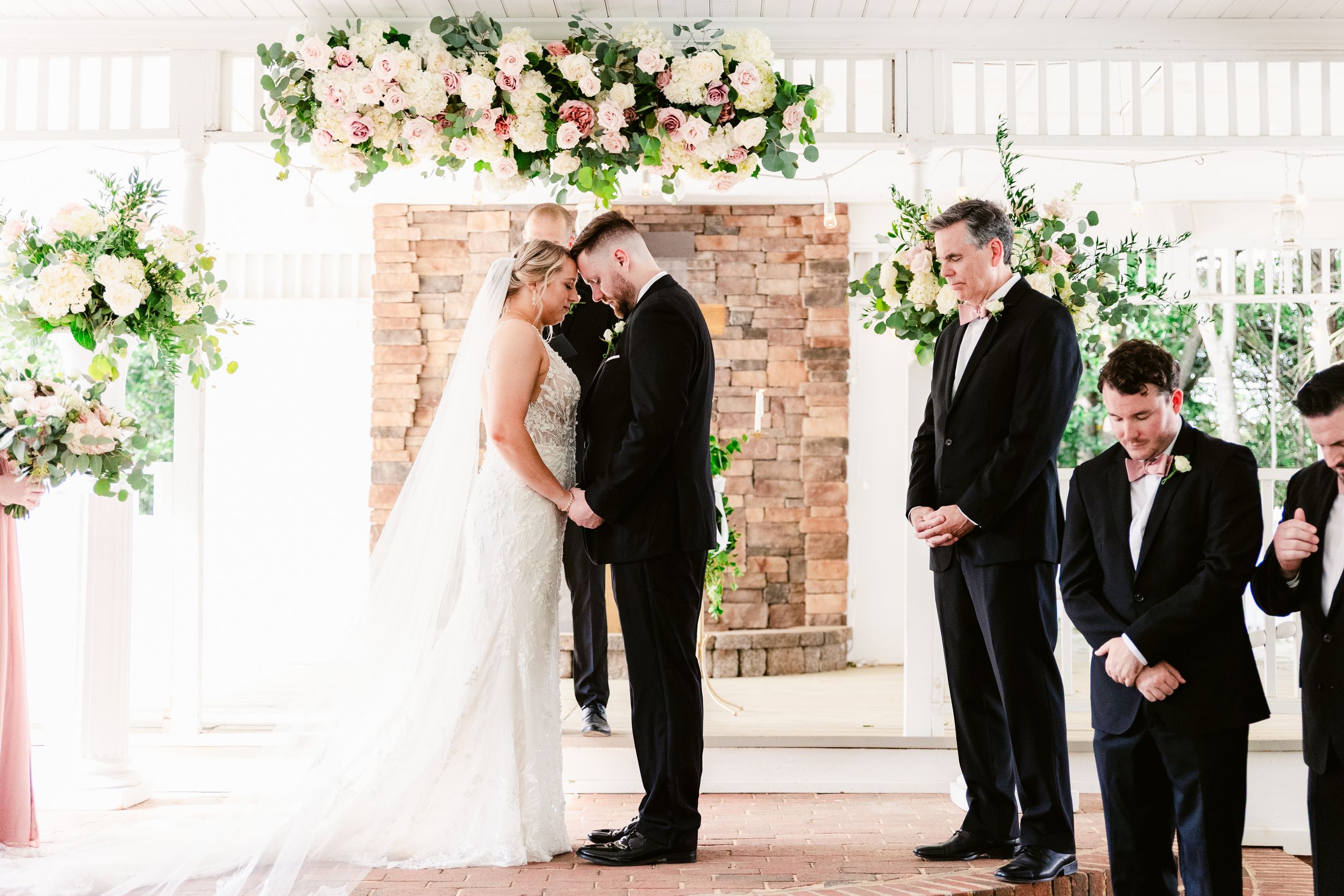 Wedding ceremony in progress under a floral arch with brick backdrop and white covered porch.