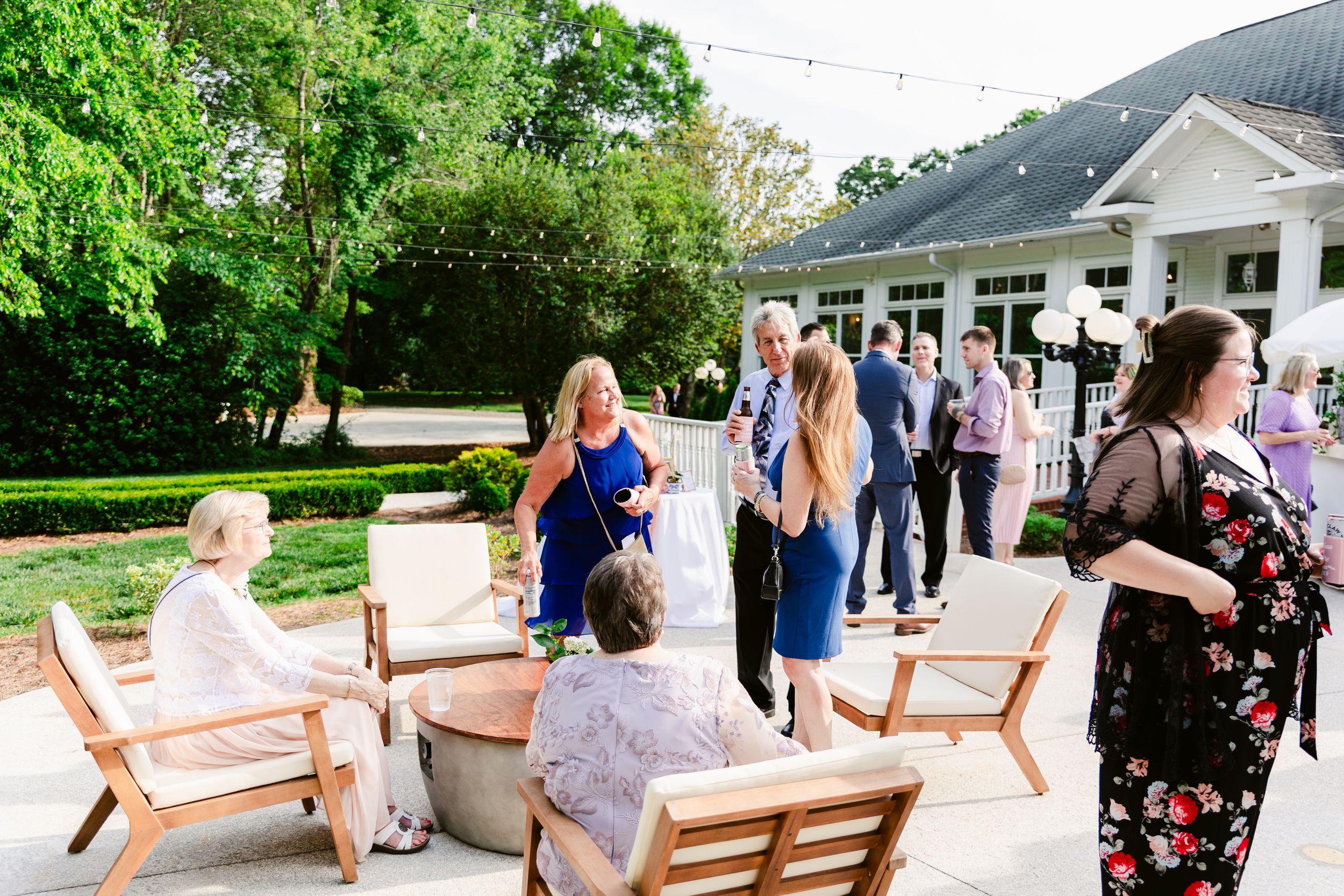 People gather on a white porch with wooden chairs during a social event at an elegant white house surrounded by greenery.
