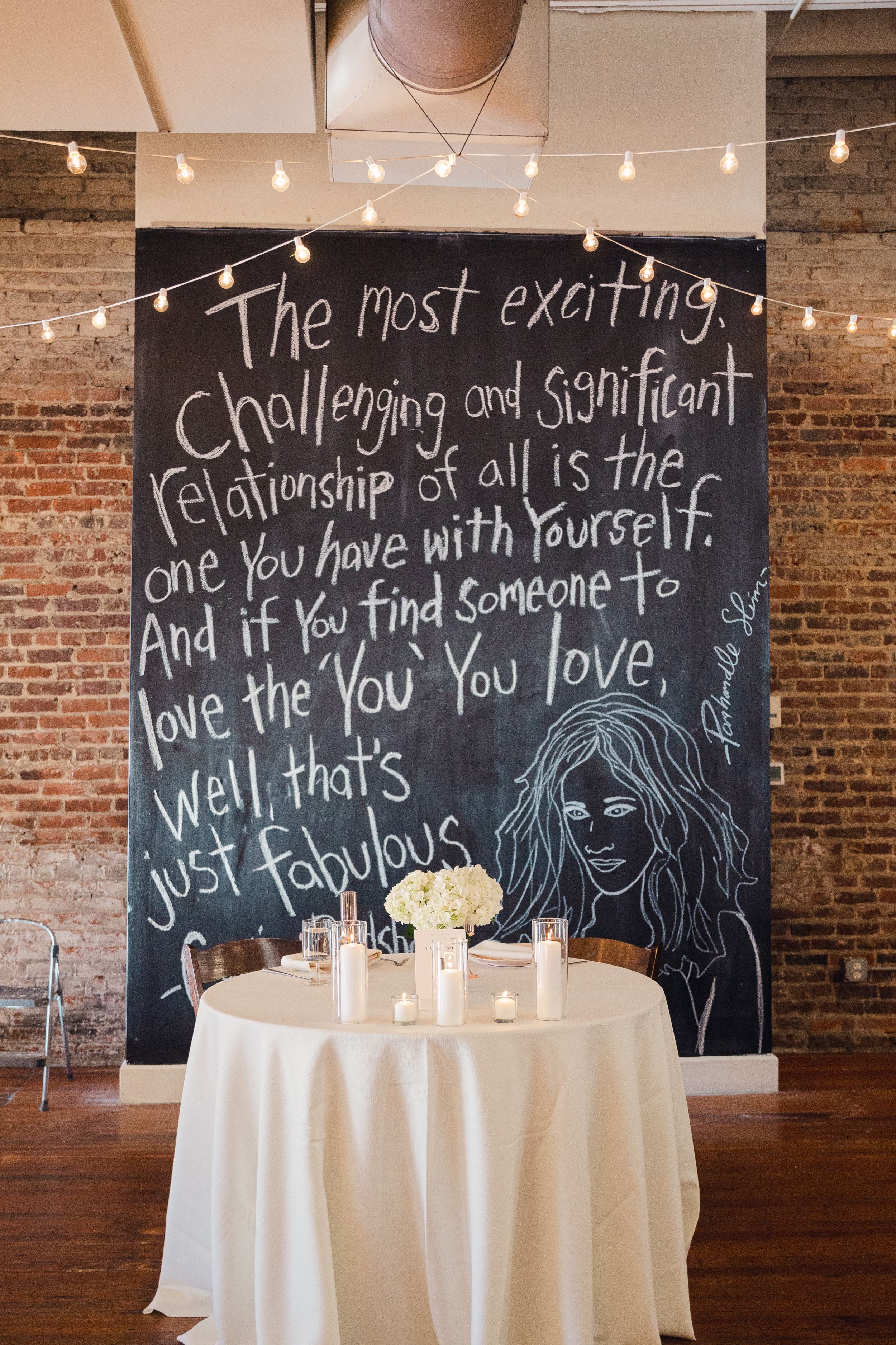 Wedding reception venue with chalkboard wall displaying inspirational quote and string lights in rustic brick setting.