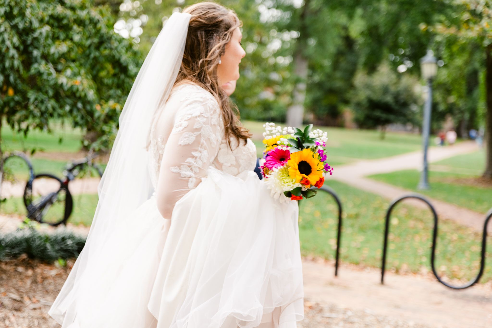 A person in a flowing white dress walks through a park pathway holding a vibrant sunflower bouquet.