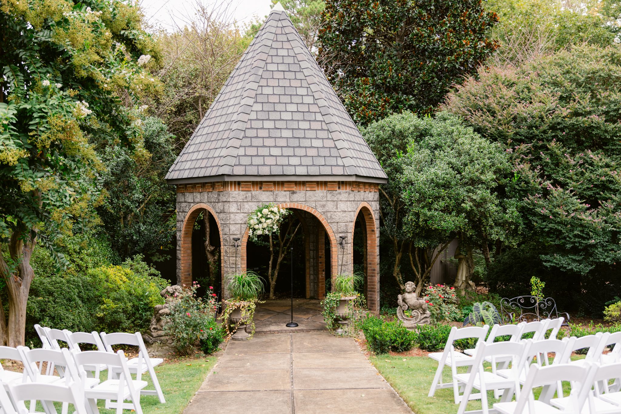 A picturesque garden wedding venue with white chairs set up in front of a stone gazebo surrounded by lush greenery.
