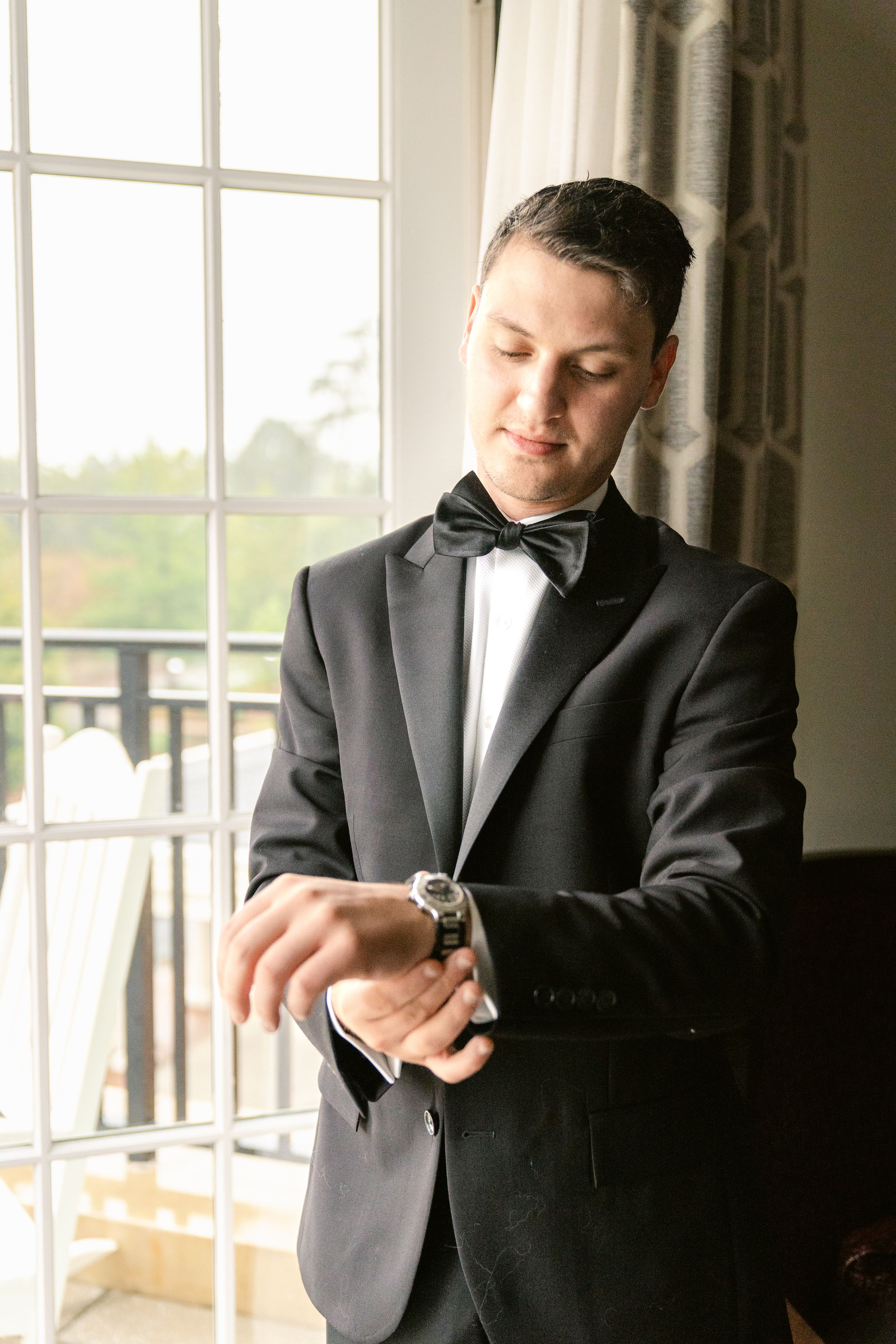 A person in a black tuxedo and bow tie checks their watch while standing by large windows in an elegant room.