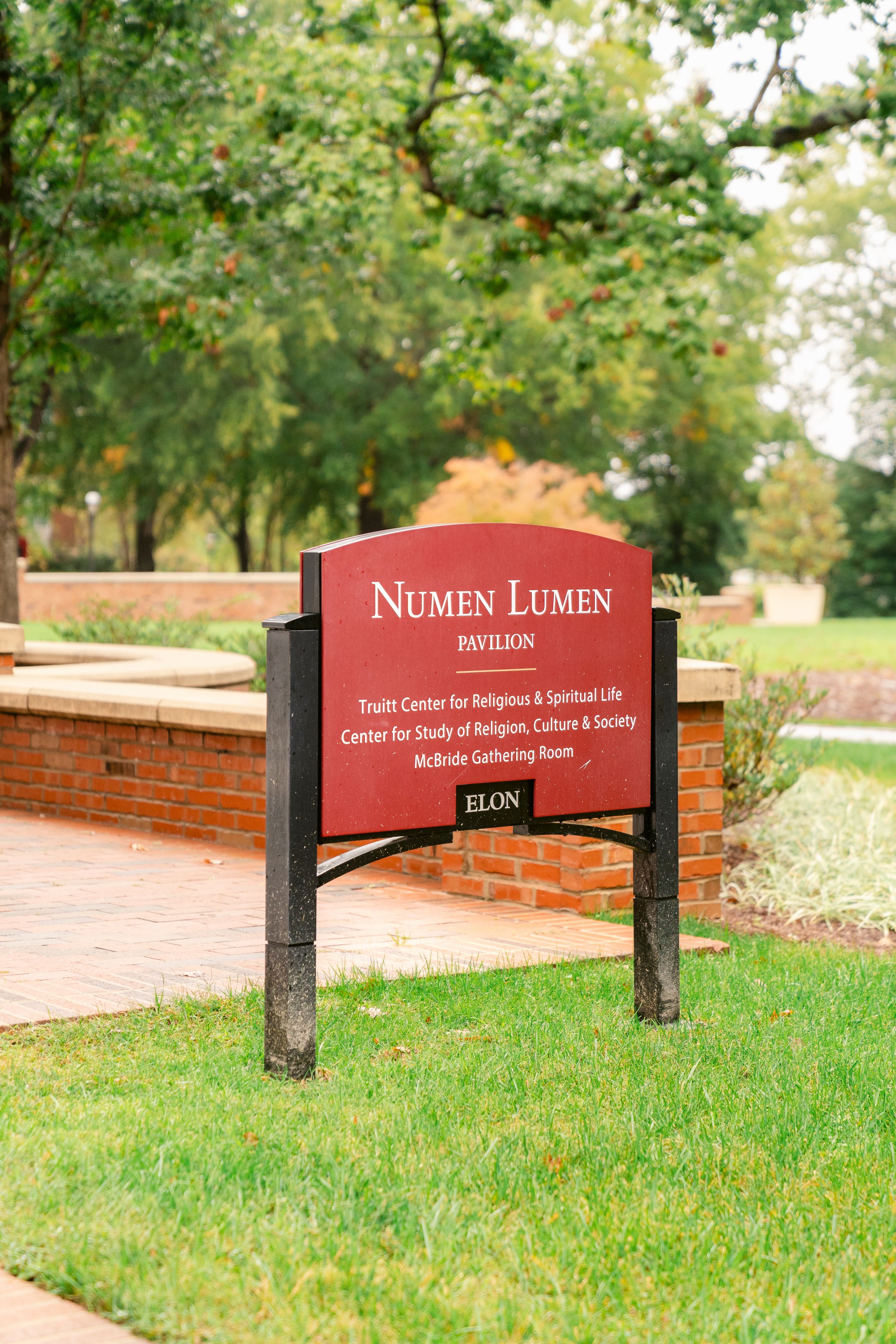 A red Numen Lumen campus sign stands in front of a green lawn and trees at Elon University.